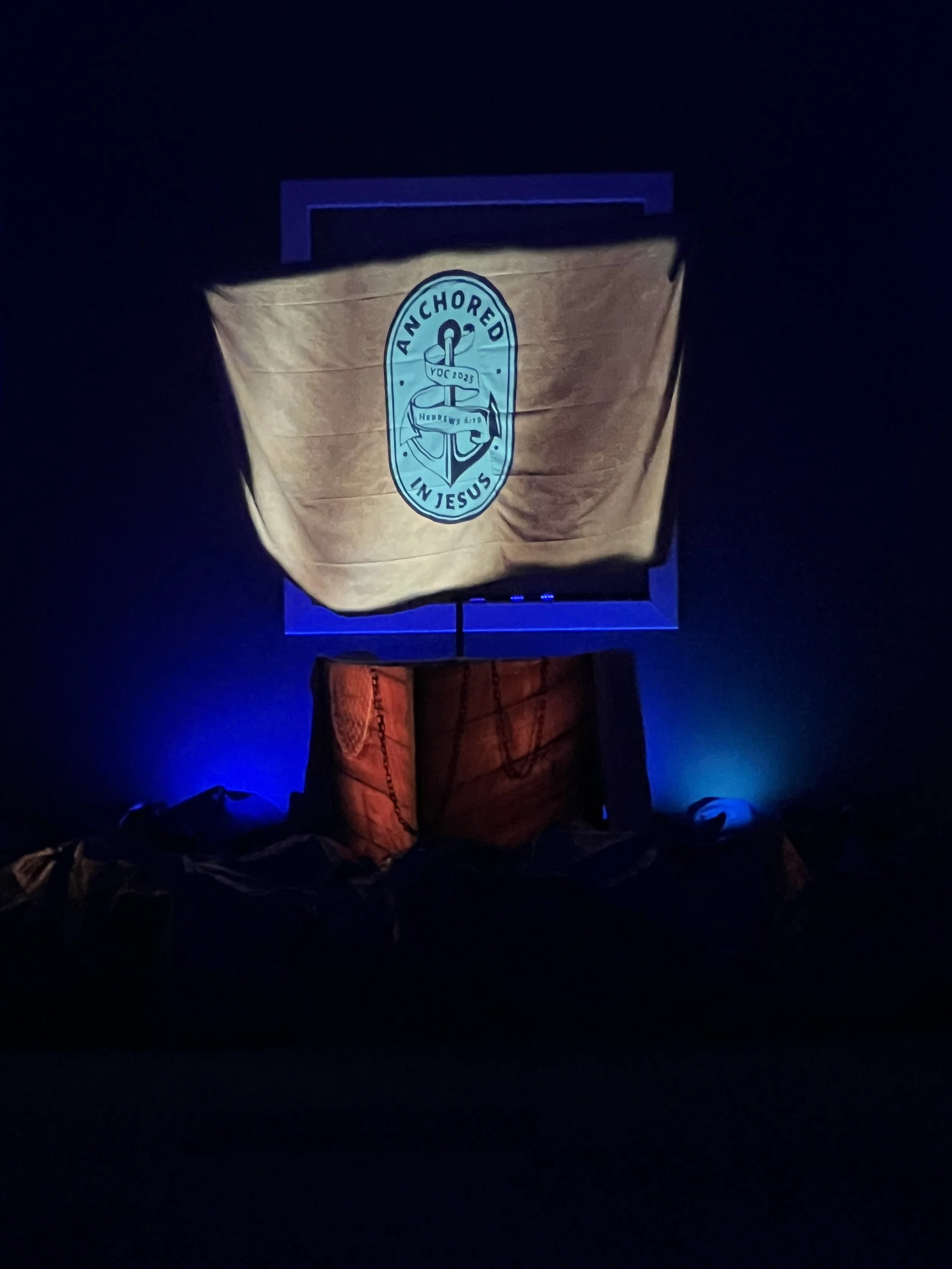A dark stage with a pirate ship set featuring an anchor and the words 'Anchored in Jesus' projected onto the sail