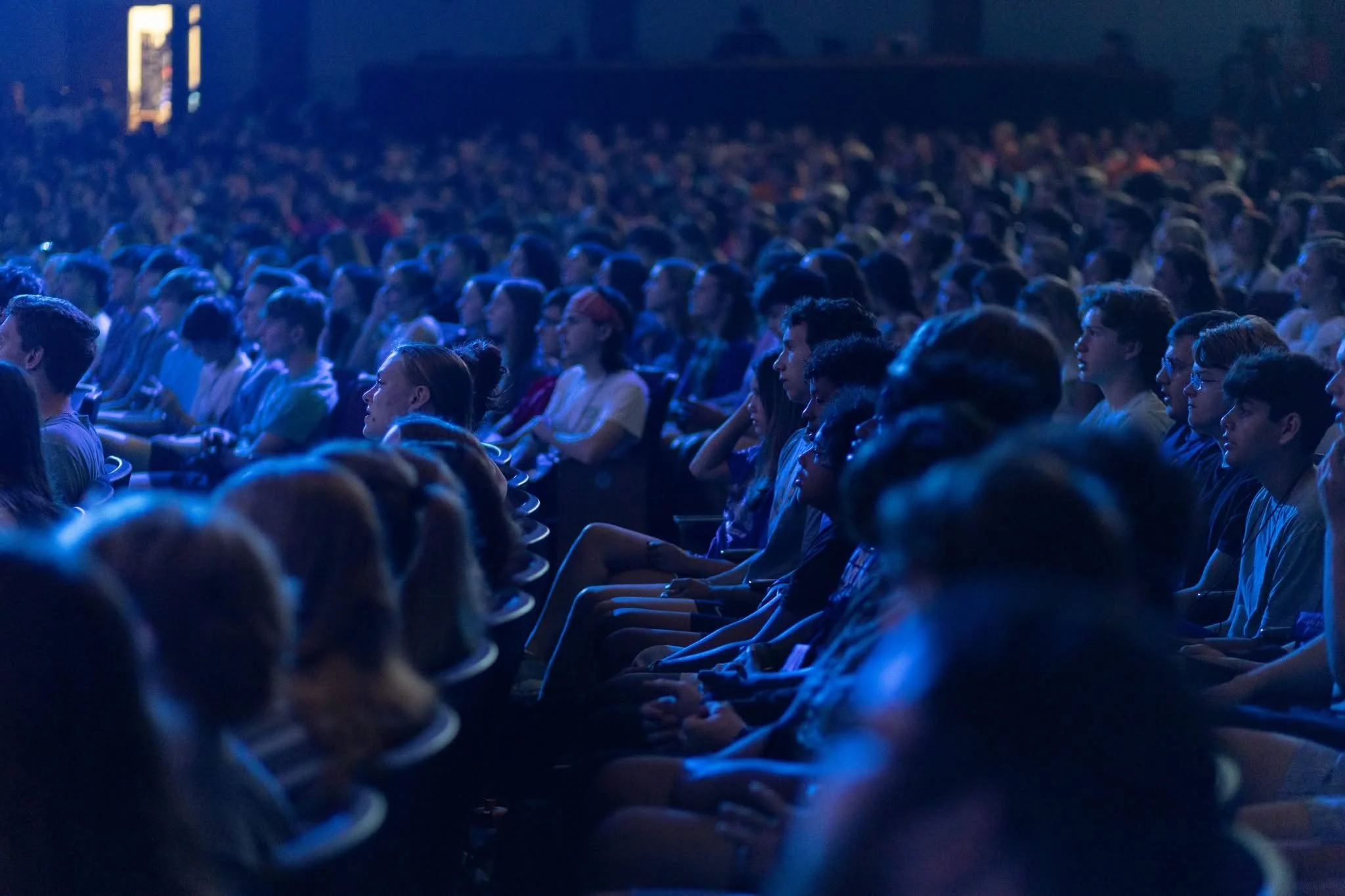 A large crowd of people seated in a dark auditorium, watching a presentation or event with dim blue lighting.