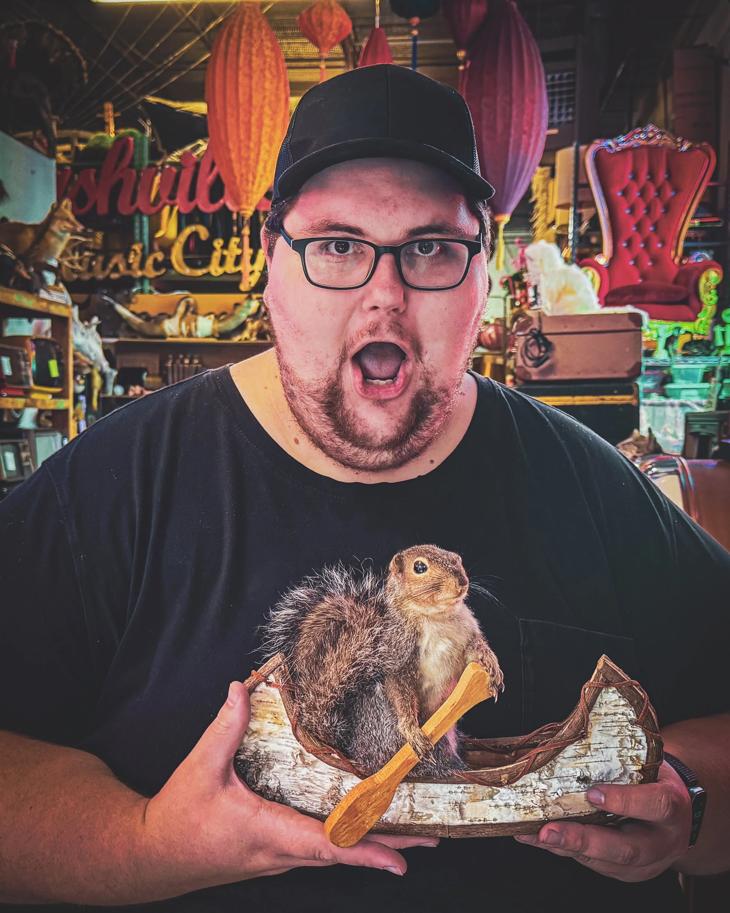A person holding a decorative canoe featuring a squirrel on a tree branch with a background of lanterns and carnival or festival decorations.