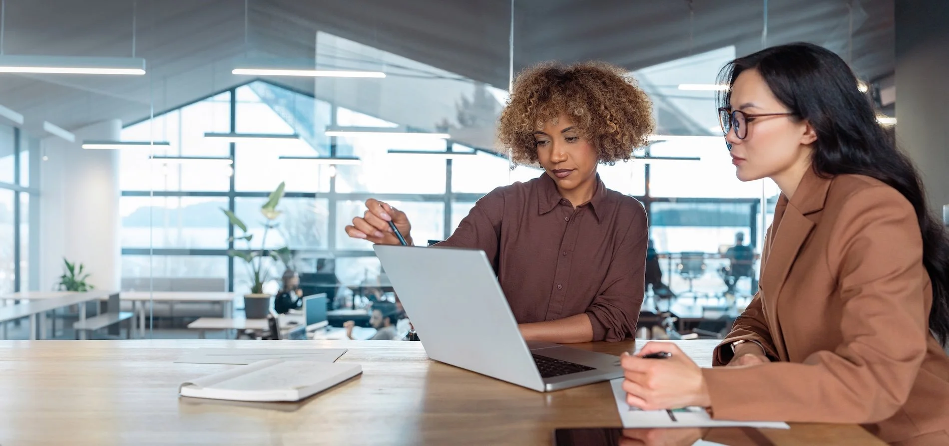 Two women sitting at a wooden table in a modern office, looking at a laptop. One woman has curly hair and is holding a pen, the other has long dark hair and glasses. Large windows and office plants are visible in the background.
