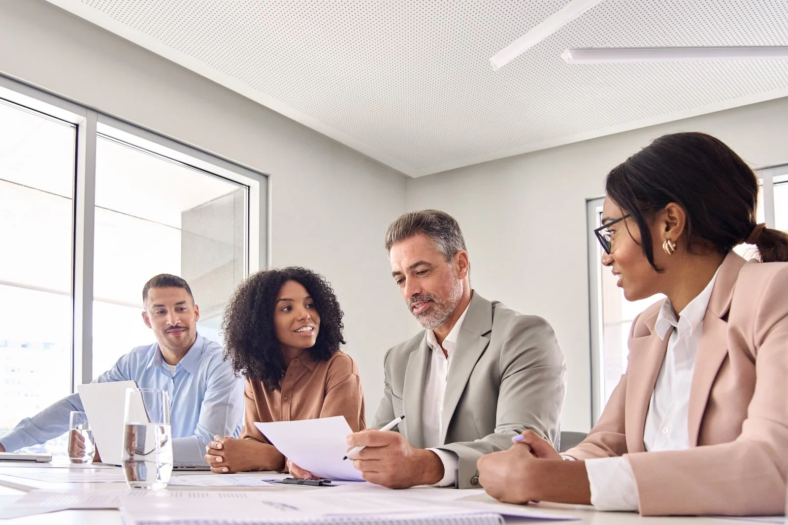 Four diverse professionals sitting at a conference table in a modern office, engaged in a discussion, with documents and glasses of water in front of them.
