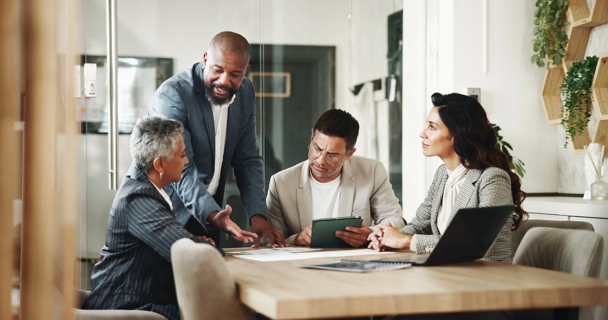 A diverse group of five businesspeople, including two women and three men, gathered around a table in a modern office, engaged in a collaborative discussion with one person showing something on a tablet.