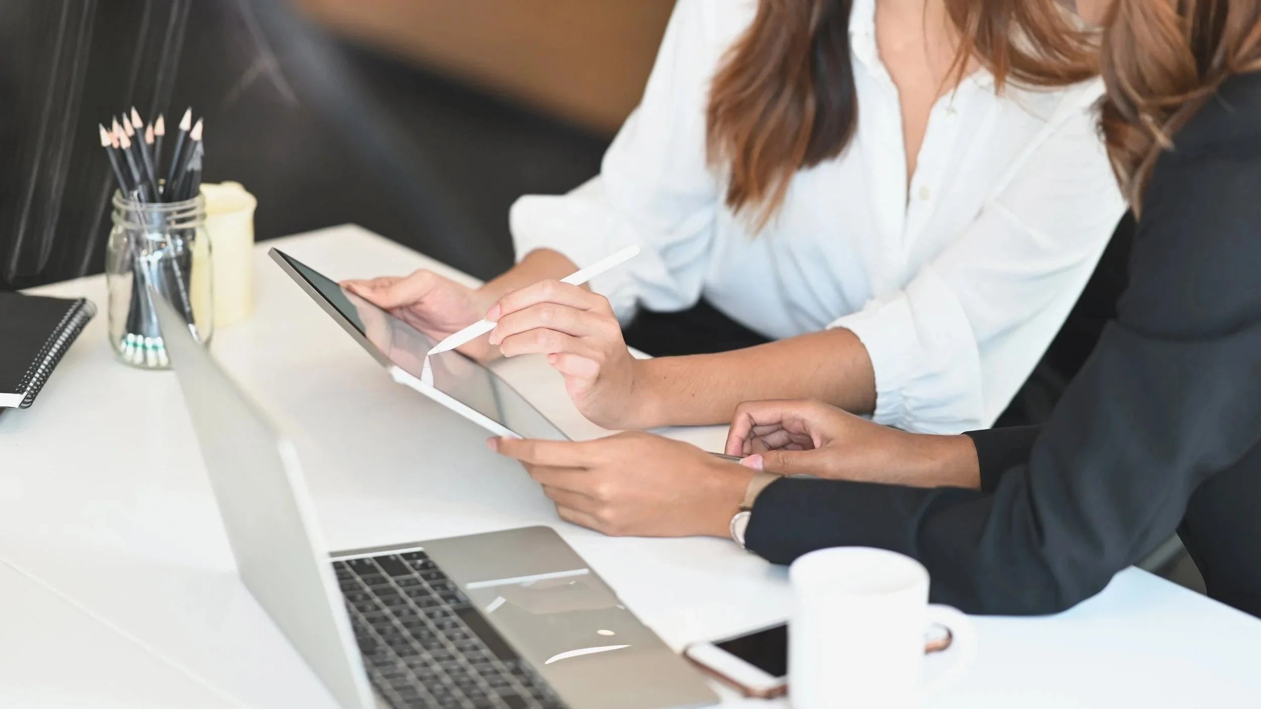 Two women sitting at a desk, one using a digital tablet with a stylus, in an office setting with a laptop, notepad, and container of pens on the desk.