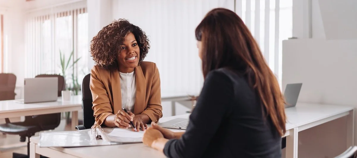 Two women having a conversation in an office, one smiling and holding a pen, the other with her back to the camera.