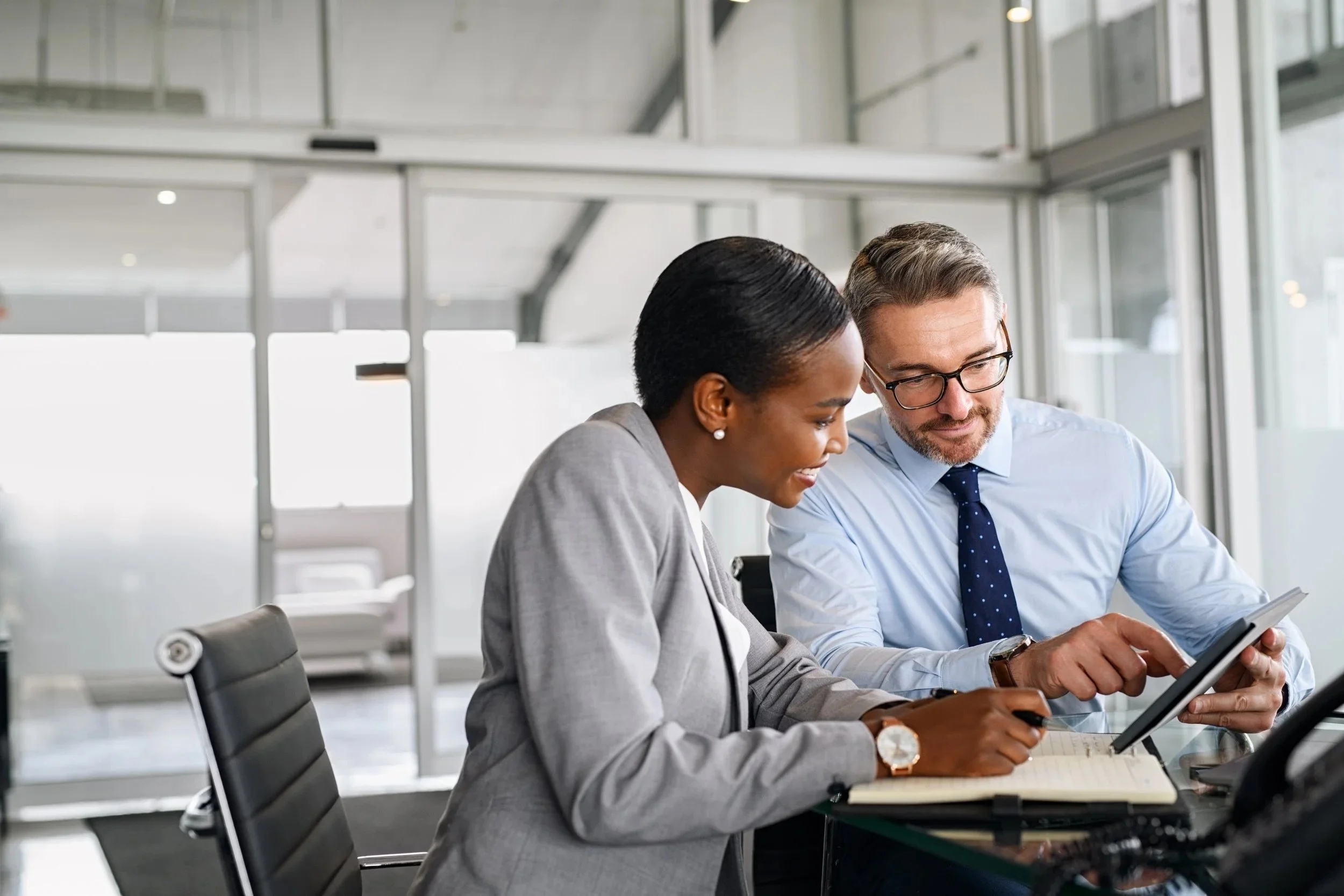 Two professionals, a woman and a man, working together and reviewing documents or digital devices in a modern office setting.