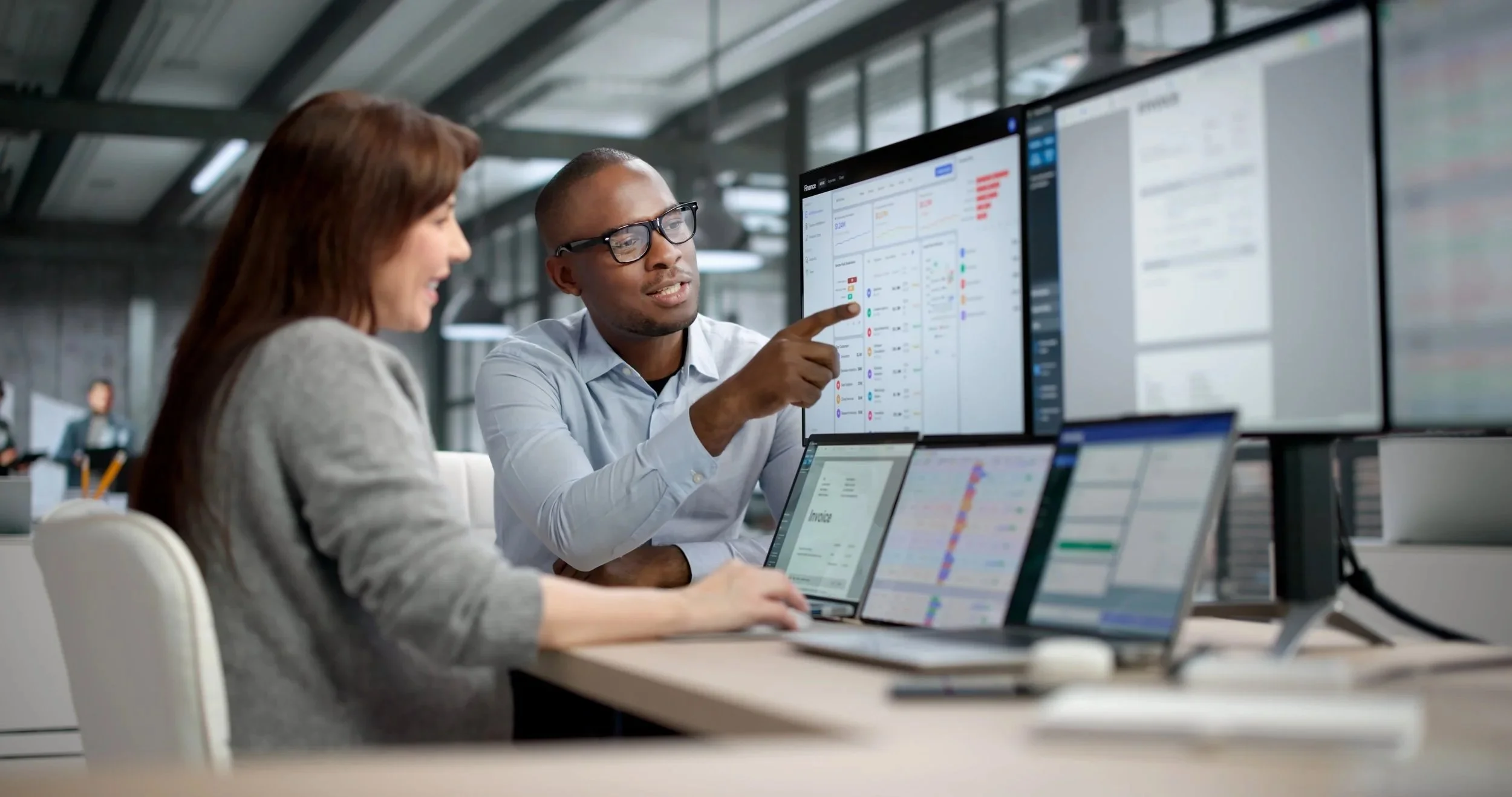 Two professionals collaborate in an office with multiple large screens displaying charts and data.