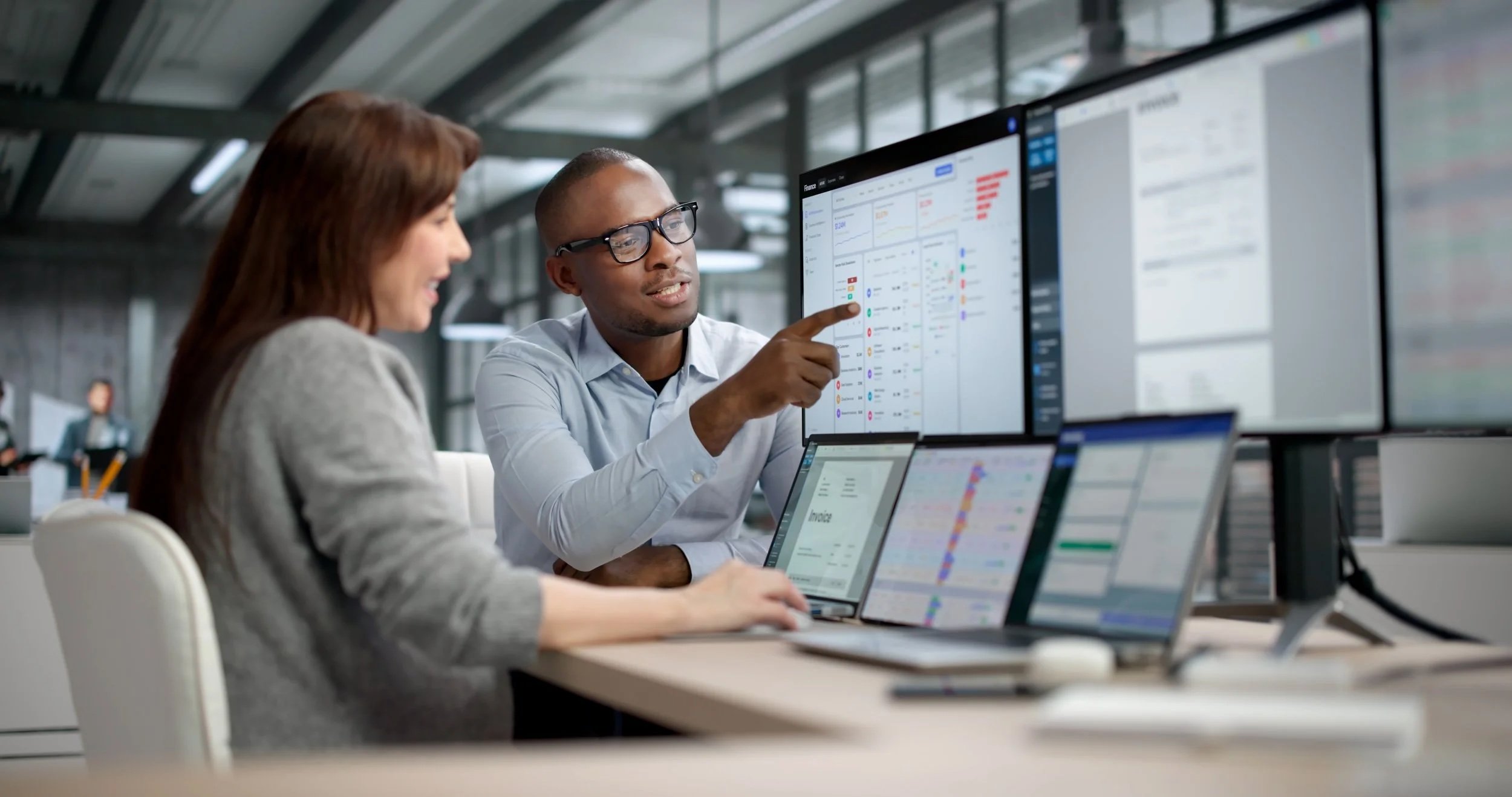 Two professionals collaborate in an office with multiple large screens displaying charts and data.