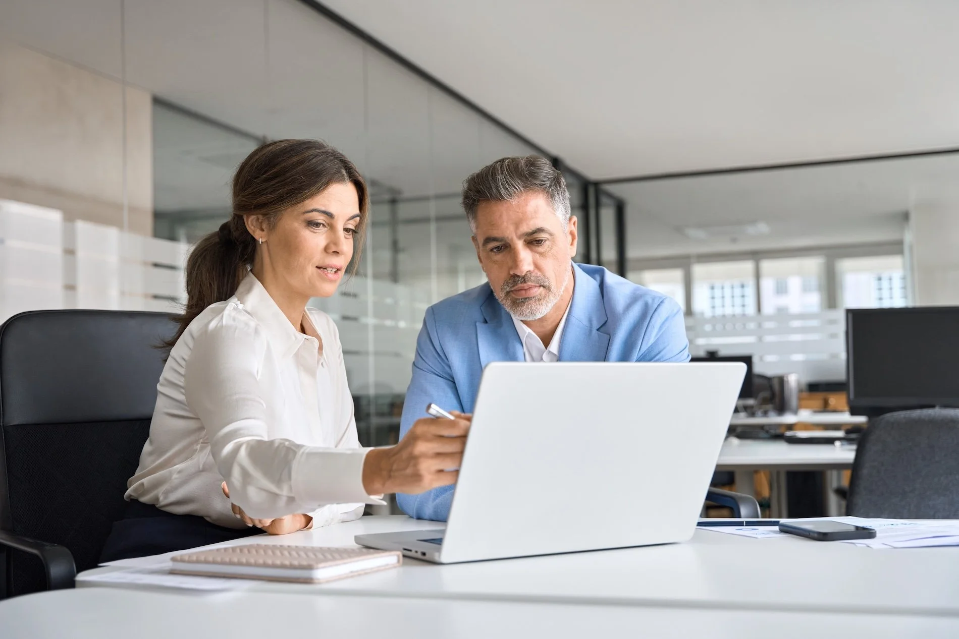 A man and woman sitting at a white office desk, looking at a laptop screen together. The woman is holding a pen and wearing a white blouse, while the man is in a blue blazer. There are notebooks and a smartphone on the desk, with an open-plan office environment in the background.