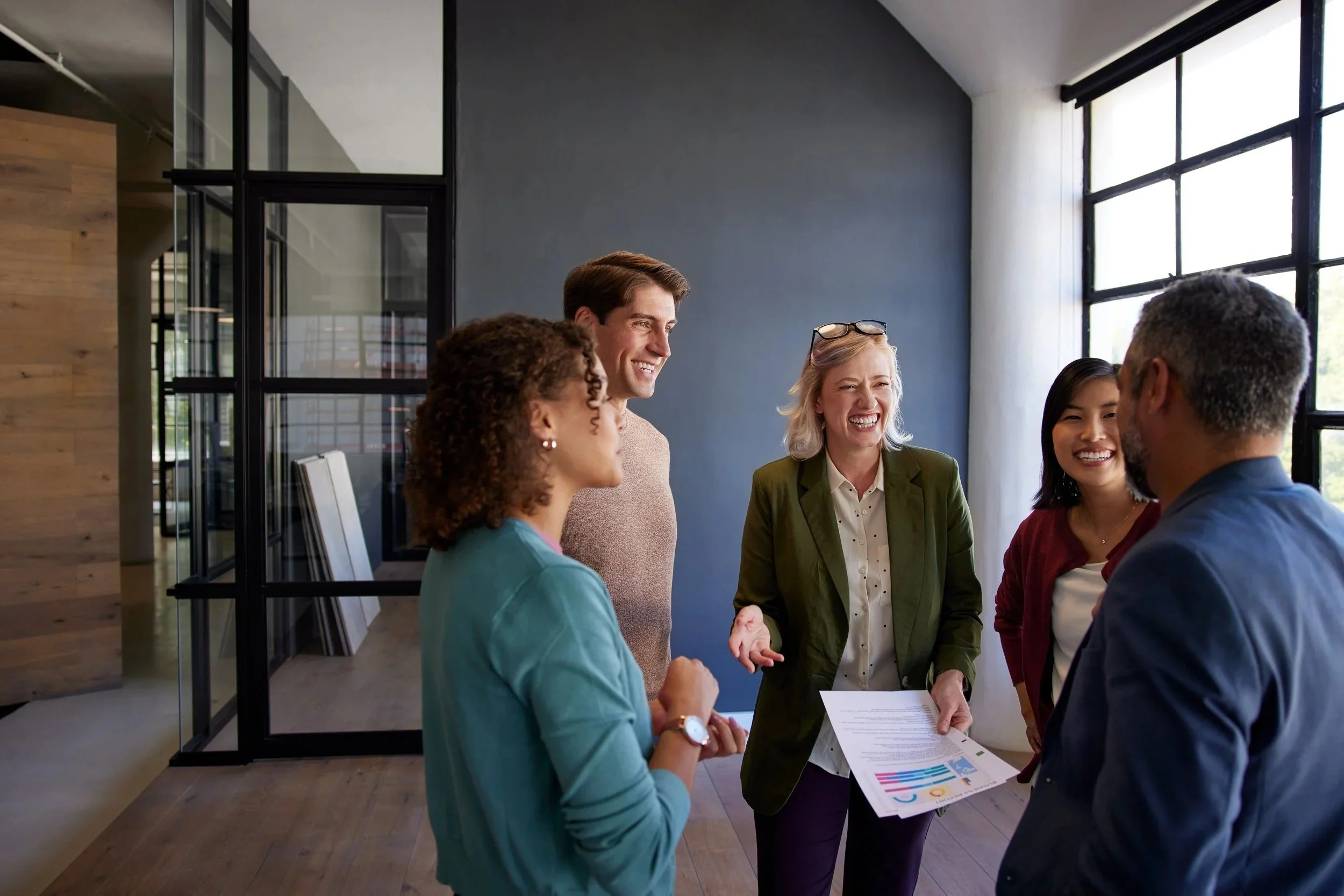 A diverse group of five people in a modern office, engaging in conversation and smiling, with one person handing a document to another.