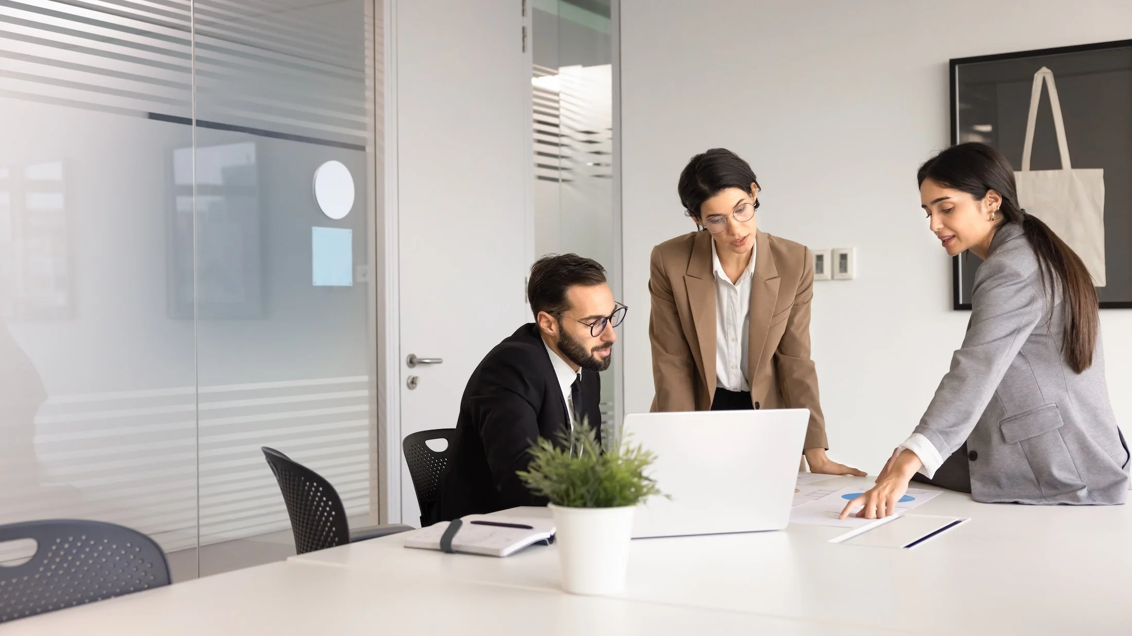 Three business professionals in a modern conference room collaborating around a laptop, with notes and a plant on the table.