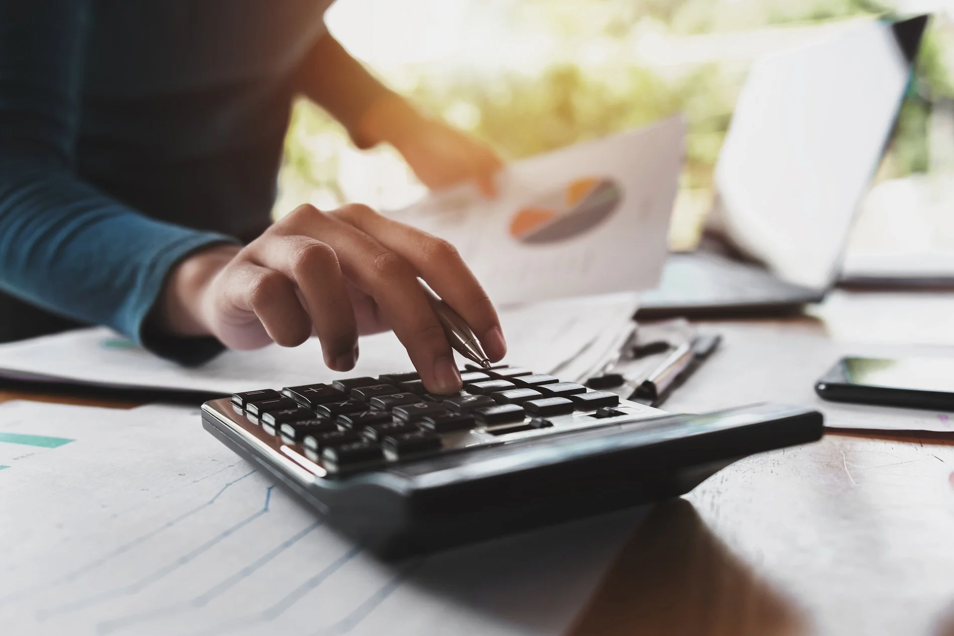 A person using a calculator, holding a pen and working with documents in a well-lit workspace.