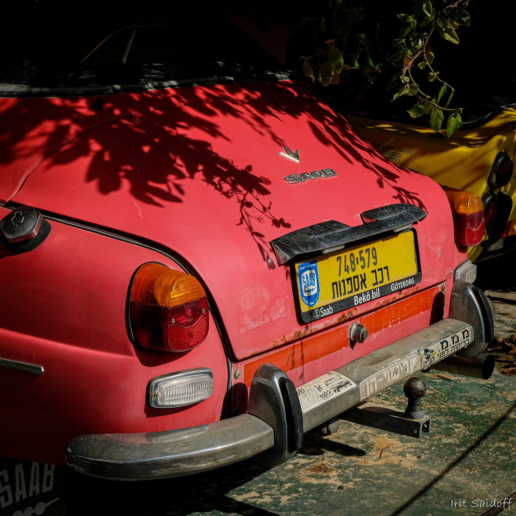 Red vehicle with a 'SAAB' badge and yellow license plate, shaded by leaves, parked on a textured ground.