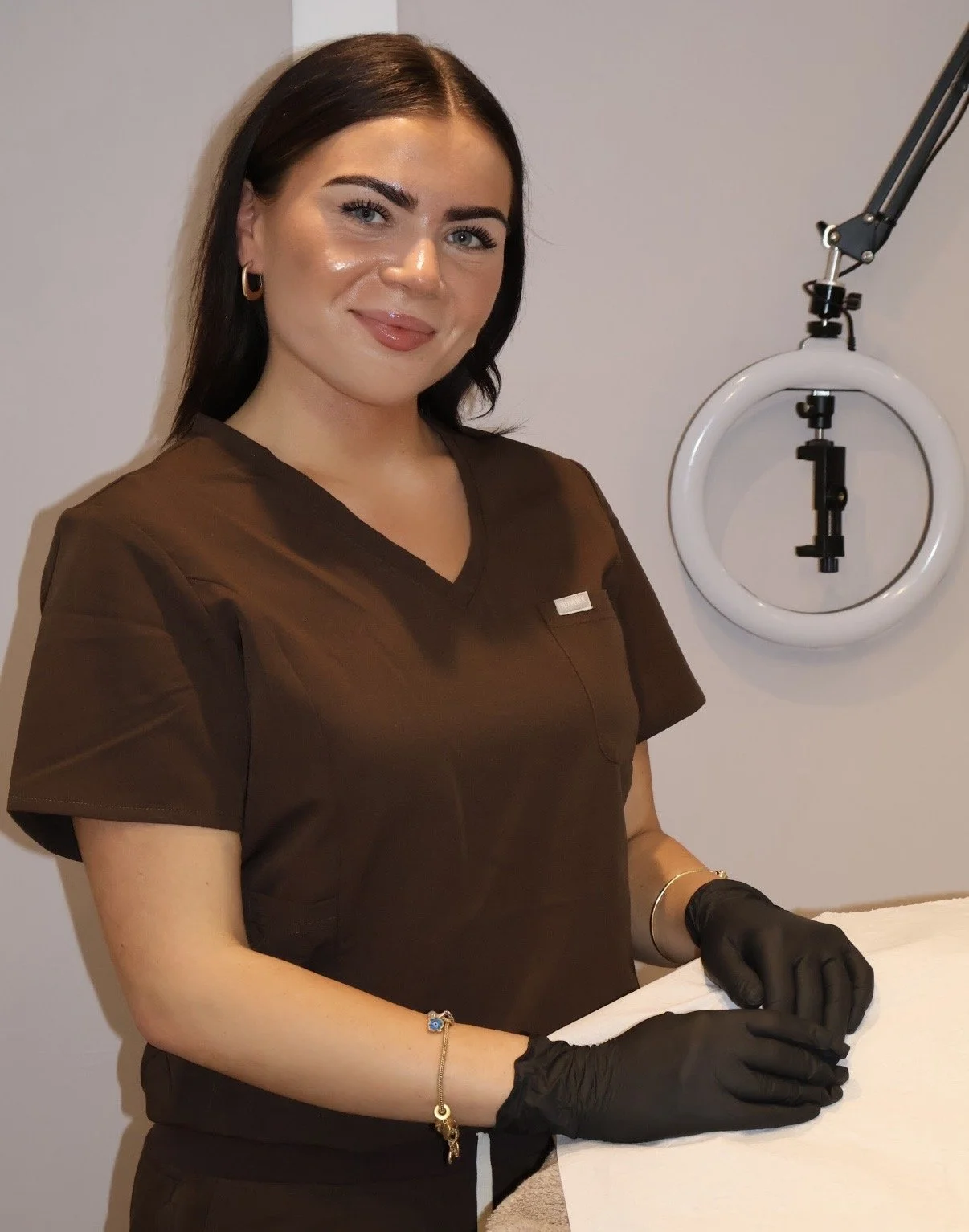 A woman with dark hair wearing medical scrubs and black gloves standing in a clinical setting with a ring light in the background.