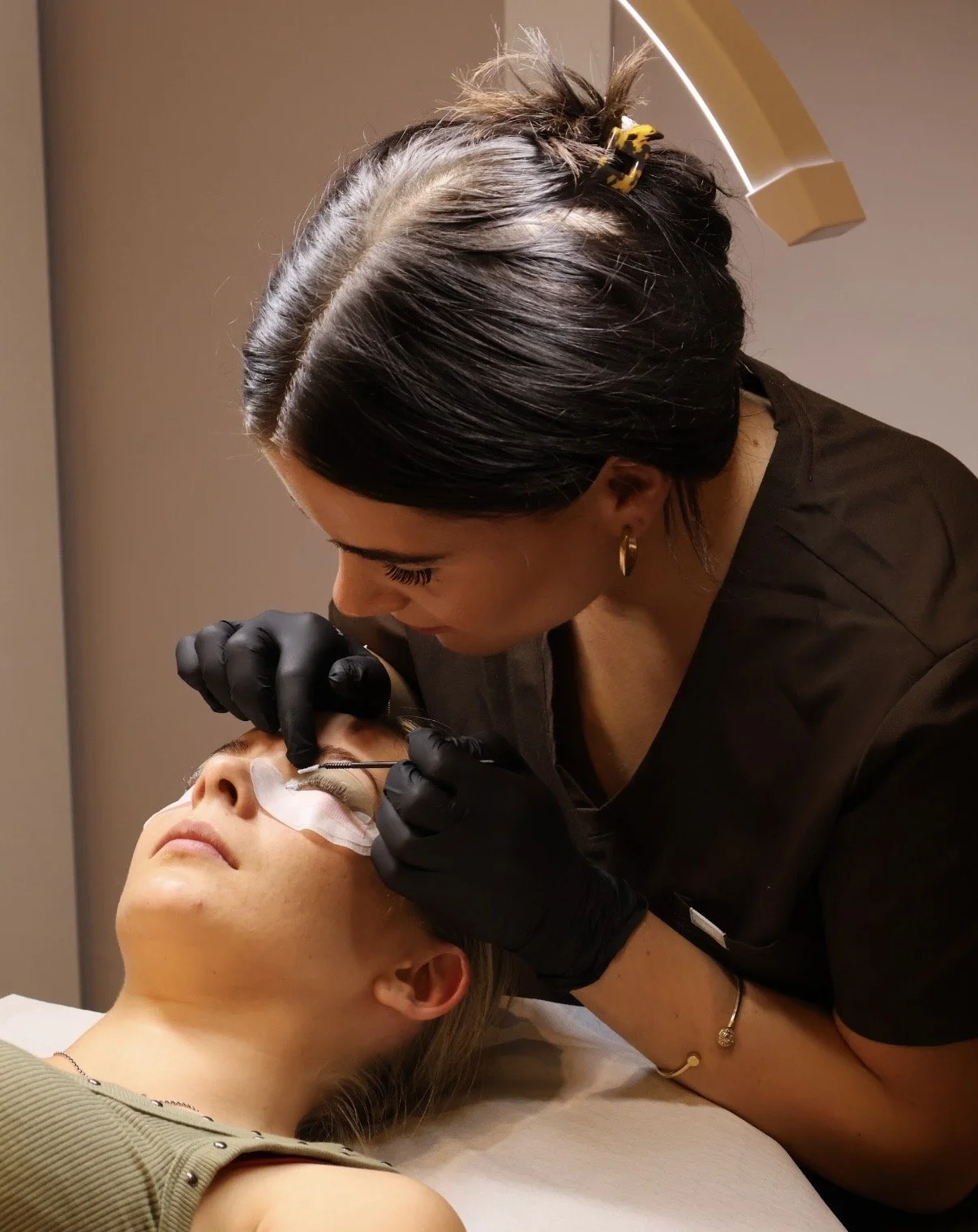 A woman lying down with her eyes closed as a technician applies eyelash extensions, wearing black gloves and working close to her eye.