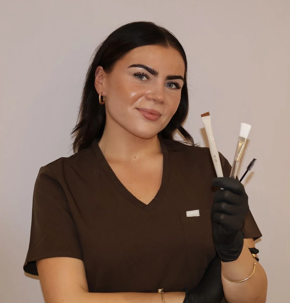 A woman with dark hair, blue eyes, and hoop earrings, dressed in a brown top, holding makeup brushes and tools, wearing black gloves, standing against a plain wall.