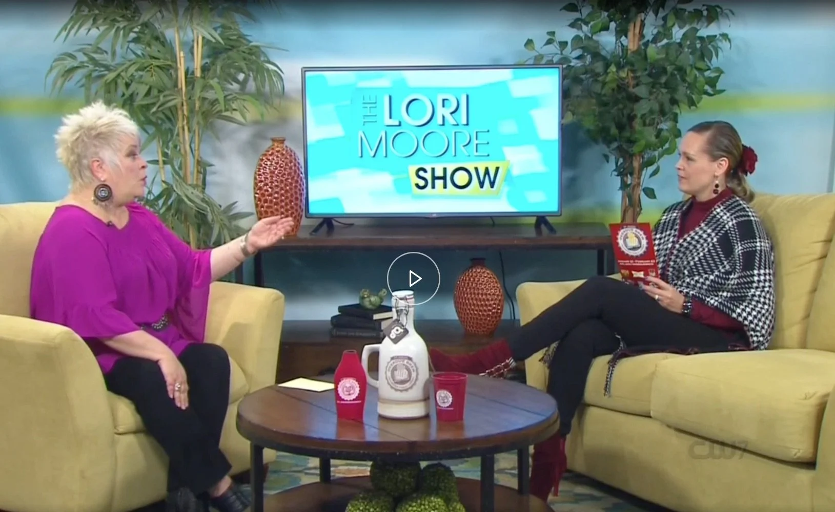 Two women talking at a table. Logo on a screen says the Lori Moore Show