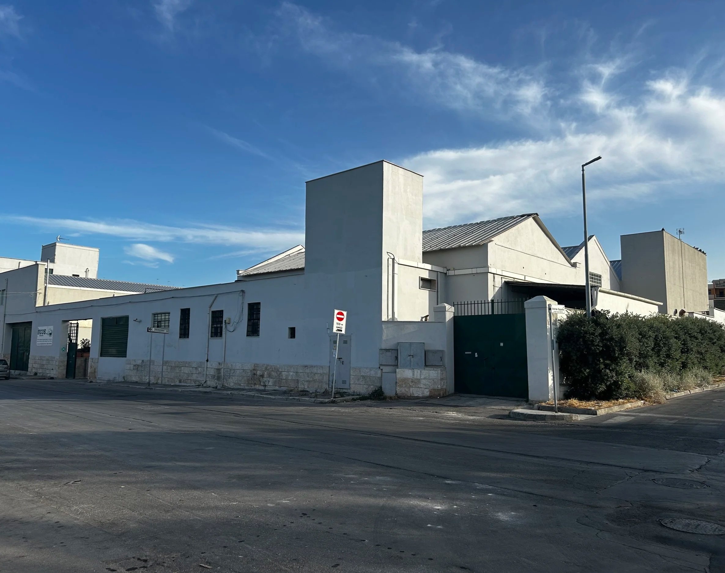 Edificio industriale bianco con tetto in metallo, lampione sulla strada e cielo azzurro con nuvole.