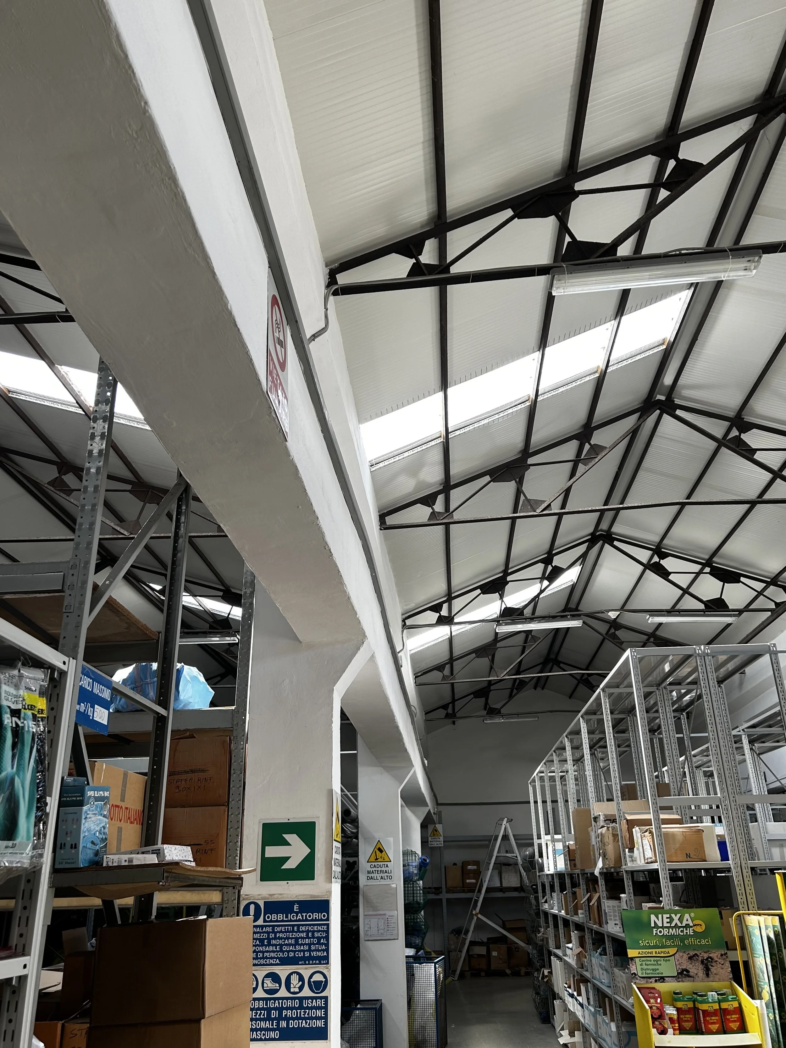 Interno di un magazzino o reparto di negozio con scaffali metallici pieni di scatole e materiali vari, pareti e soffitto con strutture in metallo, illuminazione artificiale e segnali di sicurezza e obbligo.