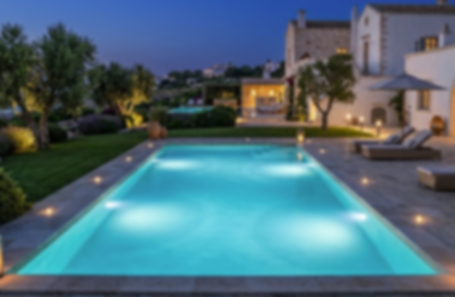 Piscina illuminata di sera in un cortile con mobili e case nello sfondo.