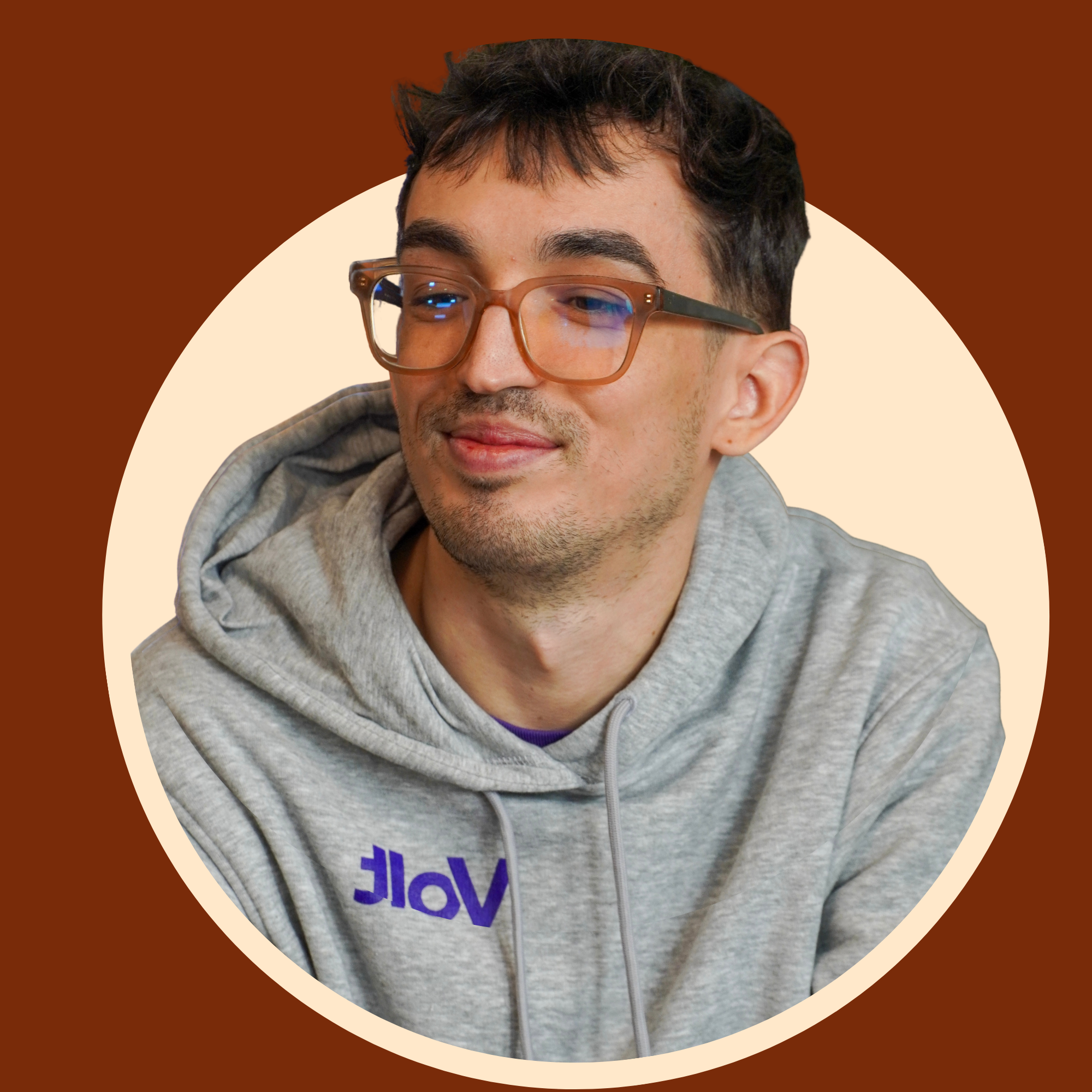 Close-up of a smiling young man with dark brown hair, wearing large brown glasses and a gray hoodie, with a background featuring layered semi-circular shapes in shades of brown and cream.