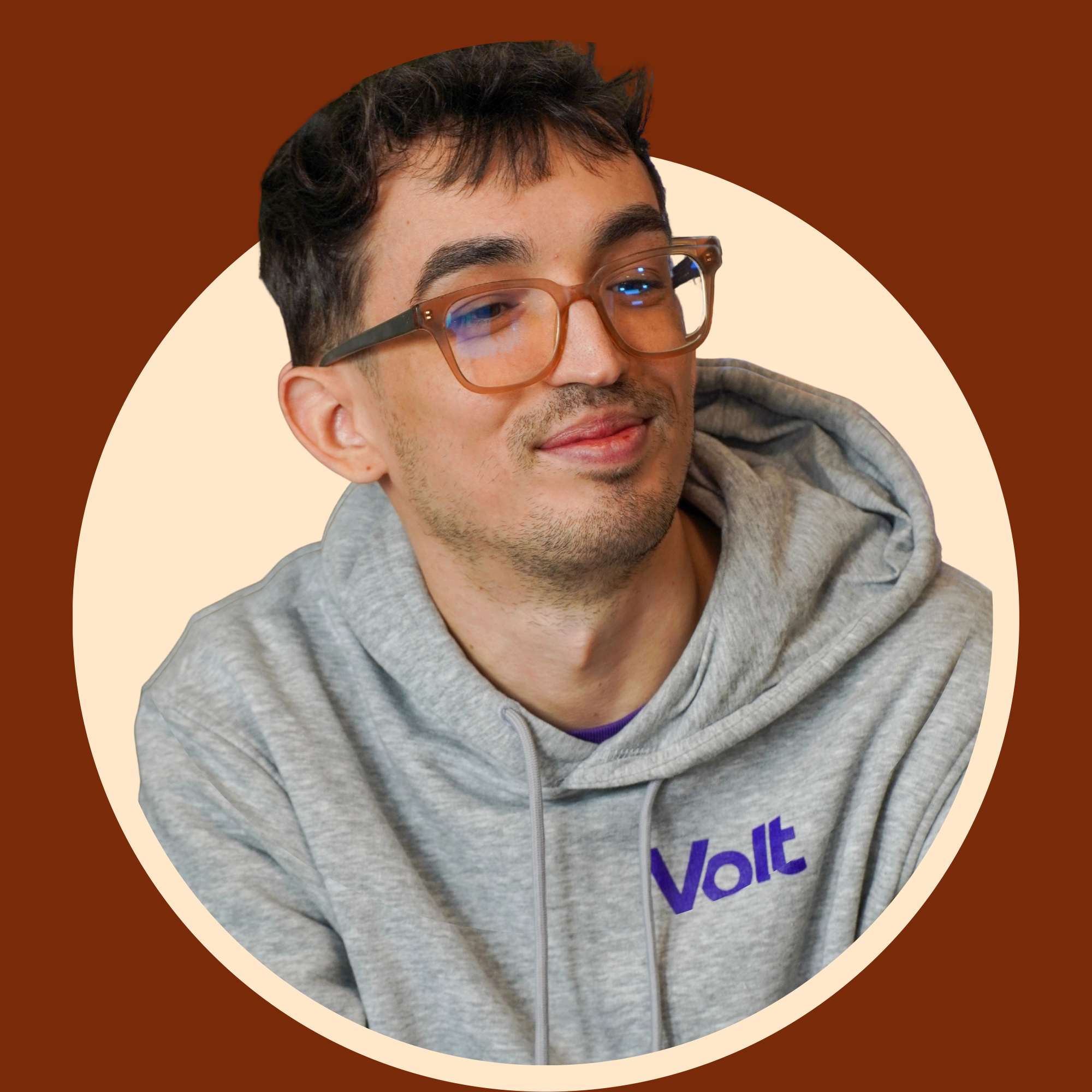 A young man with glasses and dark hair wearing a gray hoodie with purple text, smiling slightly, against a background with brown and beige curved shapes.