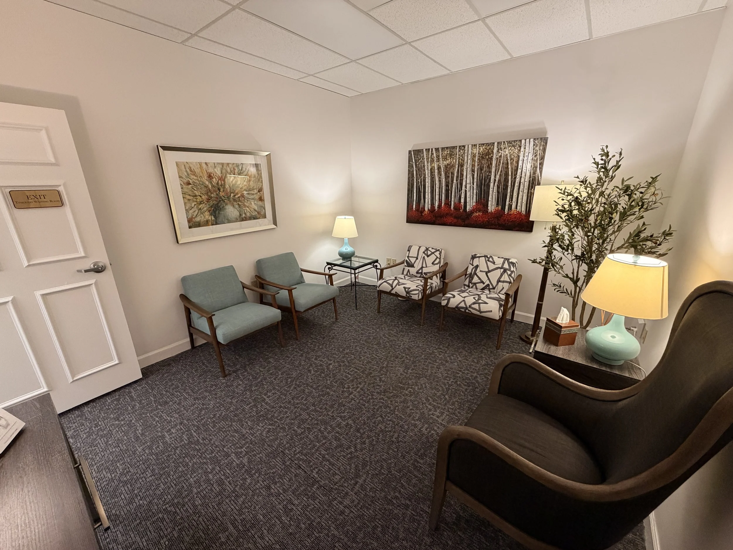 A waiting room with seats, paintings on the wall, a side table with a lamp, a plant, and an exit door.