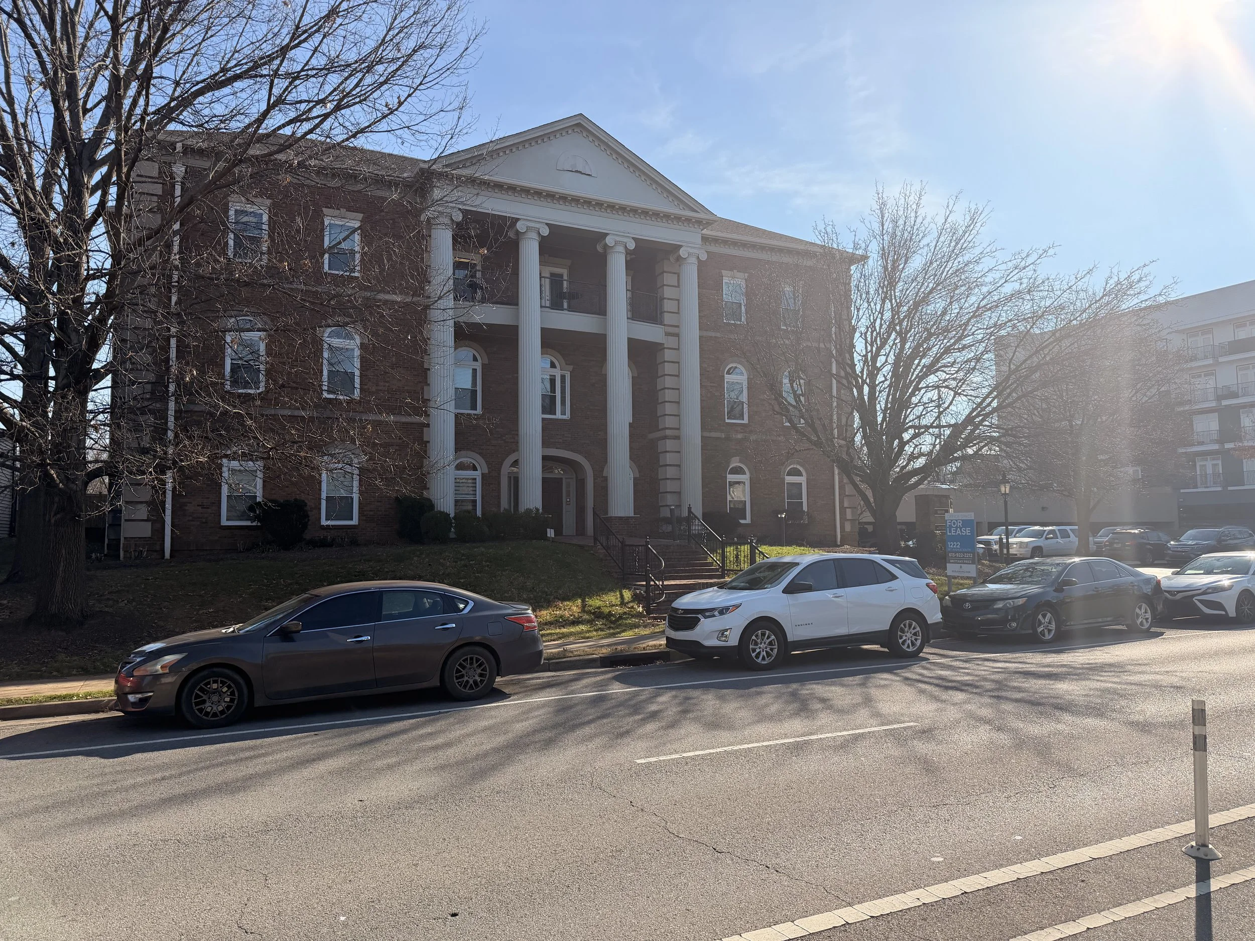 A large brick building with white columns and a triangular pediment on the front. There are trees in front of the building and a parking lot with several parked cars.