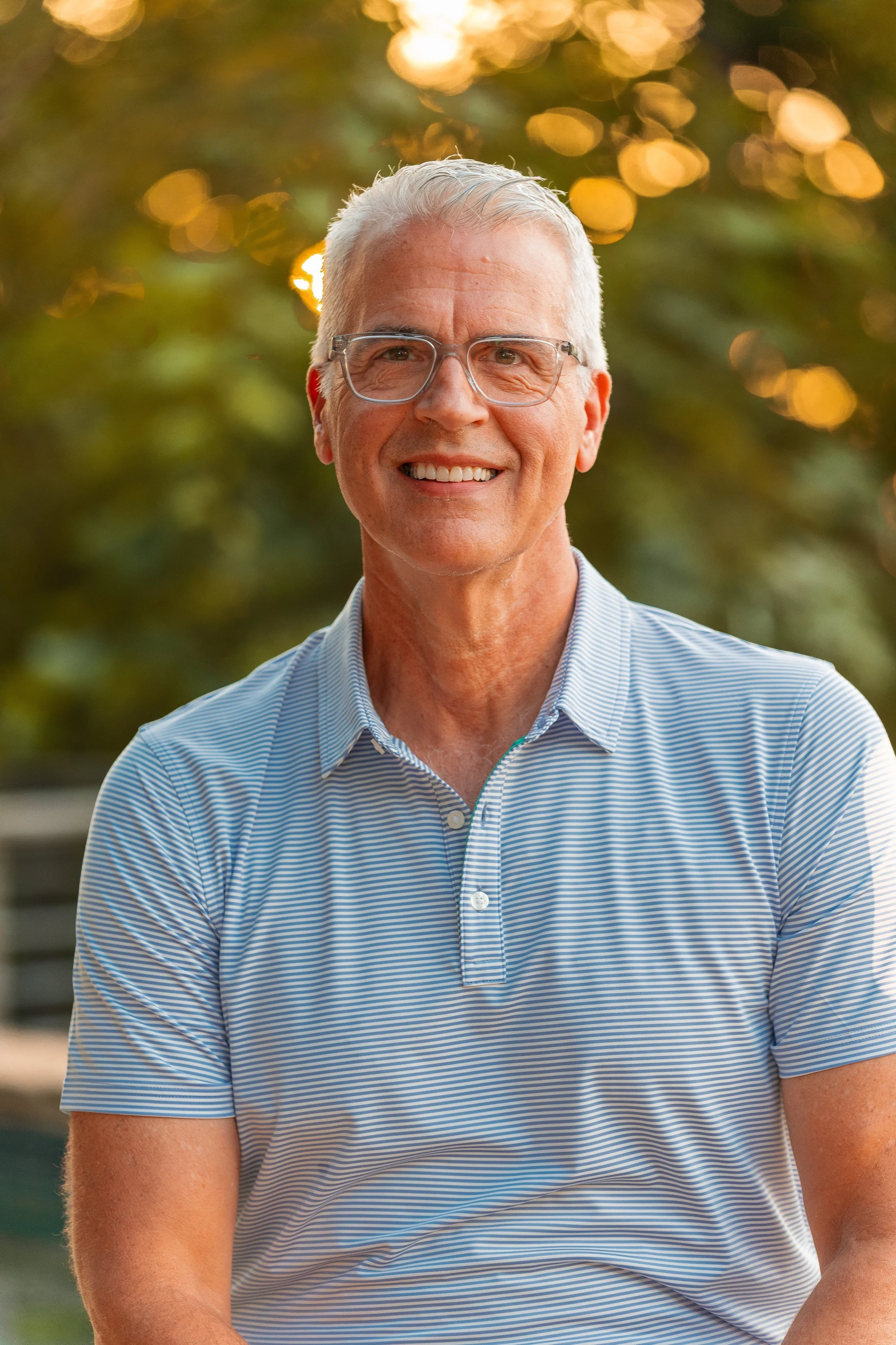 Brian Poynter smiling and wearing glasses and a blue and white striped shirt. Outdoors during sunset with blurred trees and sunlight in the background.