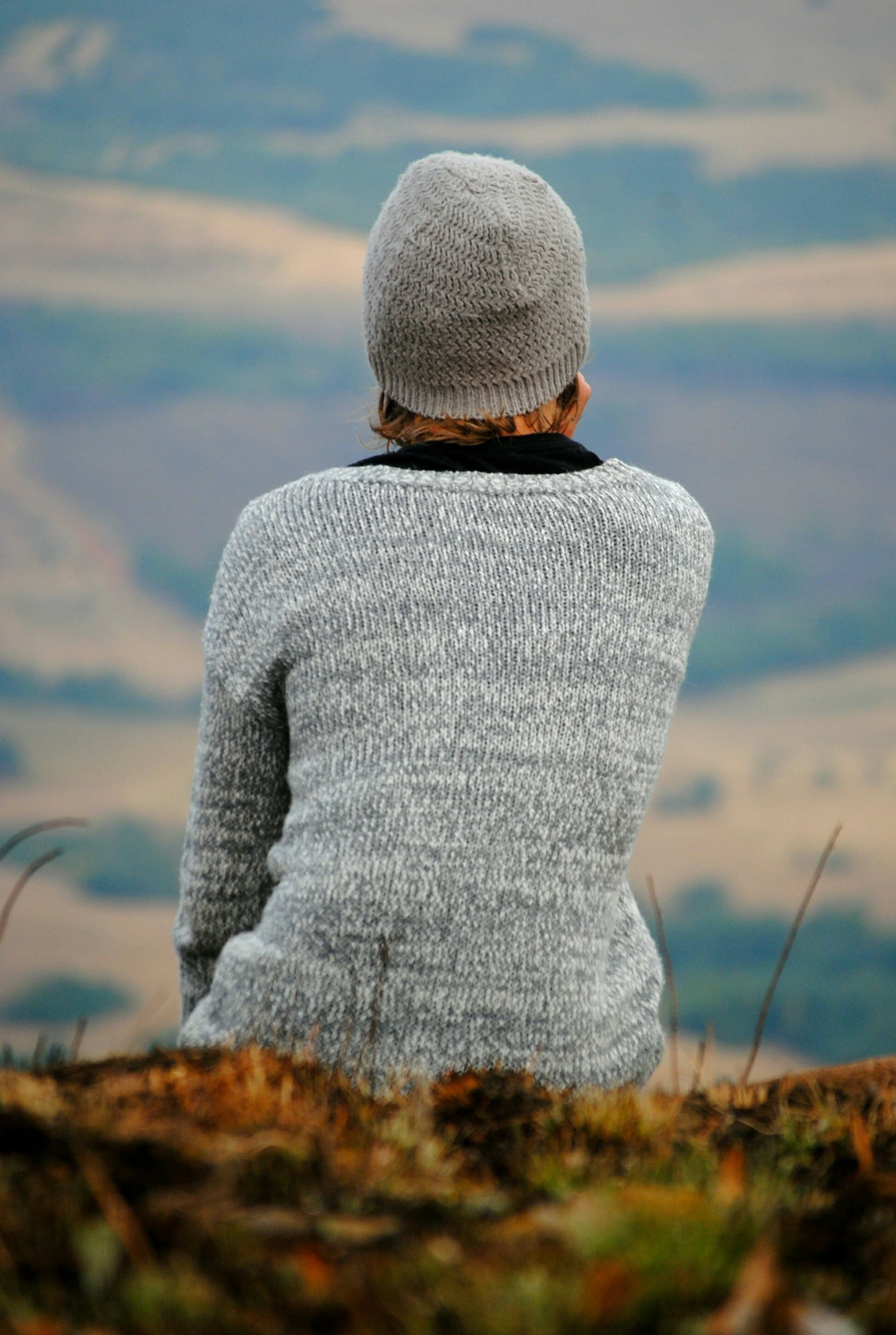 Person wearing a gray knitted beanie and gray sweater, viewed from behind, standing outdoors with a cloudy sky in the background.