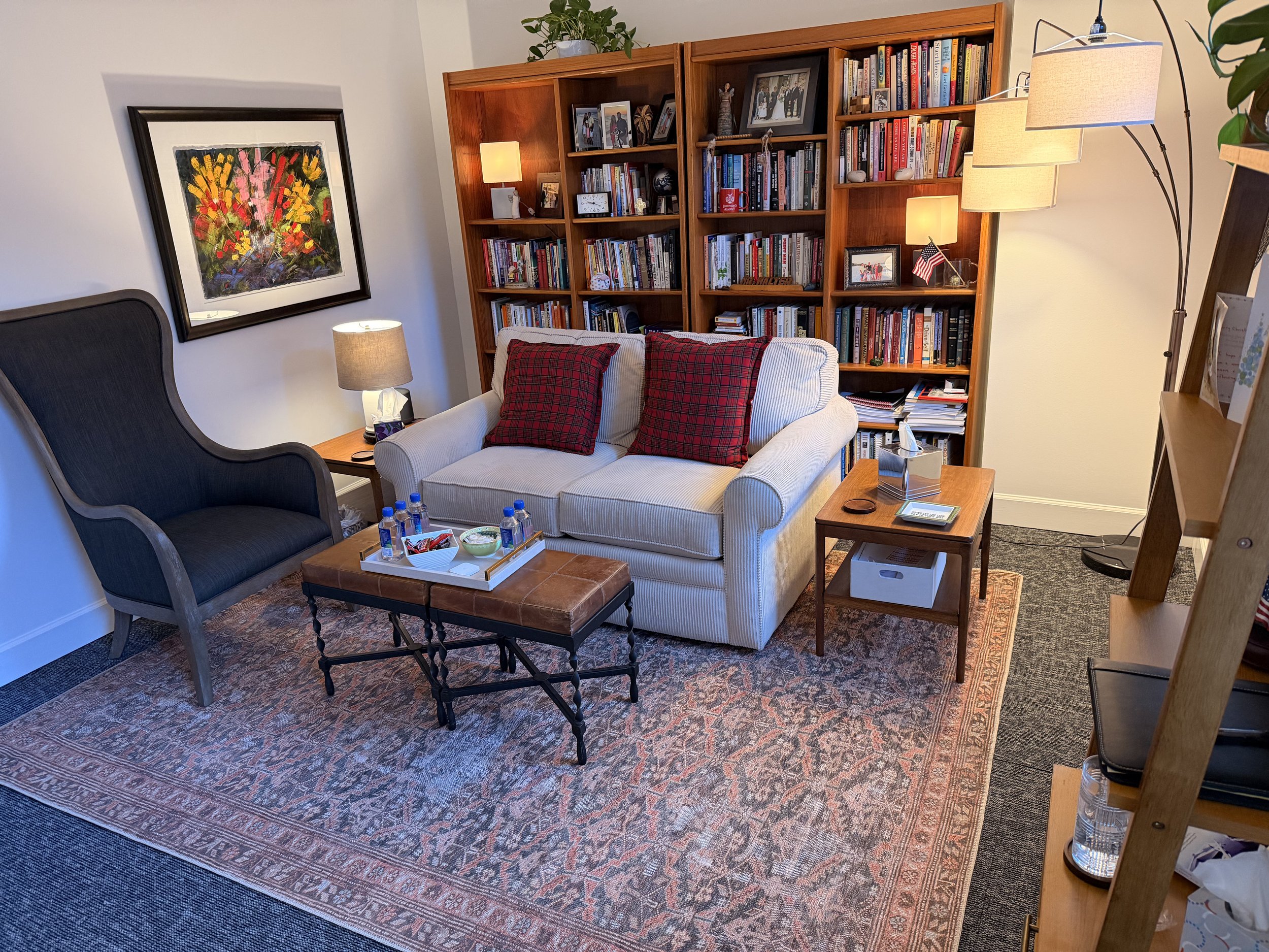 A cozy living room corner with a beige sofa with red plaid pillows, a dark armchair, a wooden bookshelf filled with books and framed photographs, a small side table with snacks and drinks, and two table lamps providing warm lighting. There is a colorful abstract painting on the wall and a patterned rug on the floor.