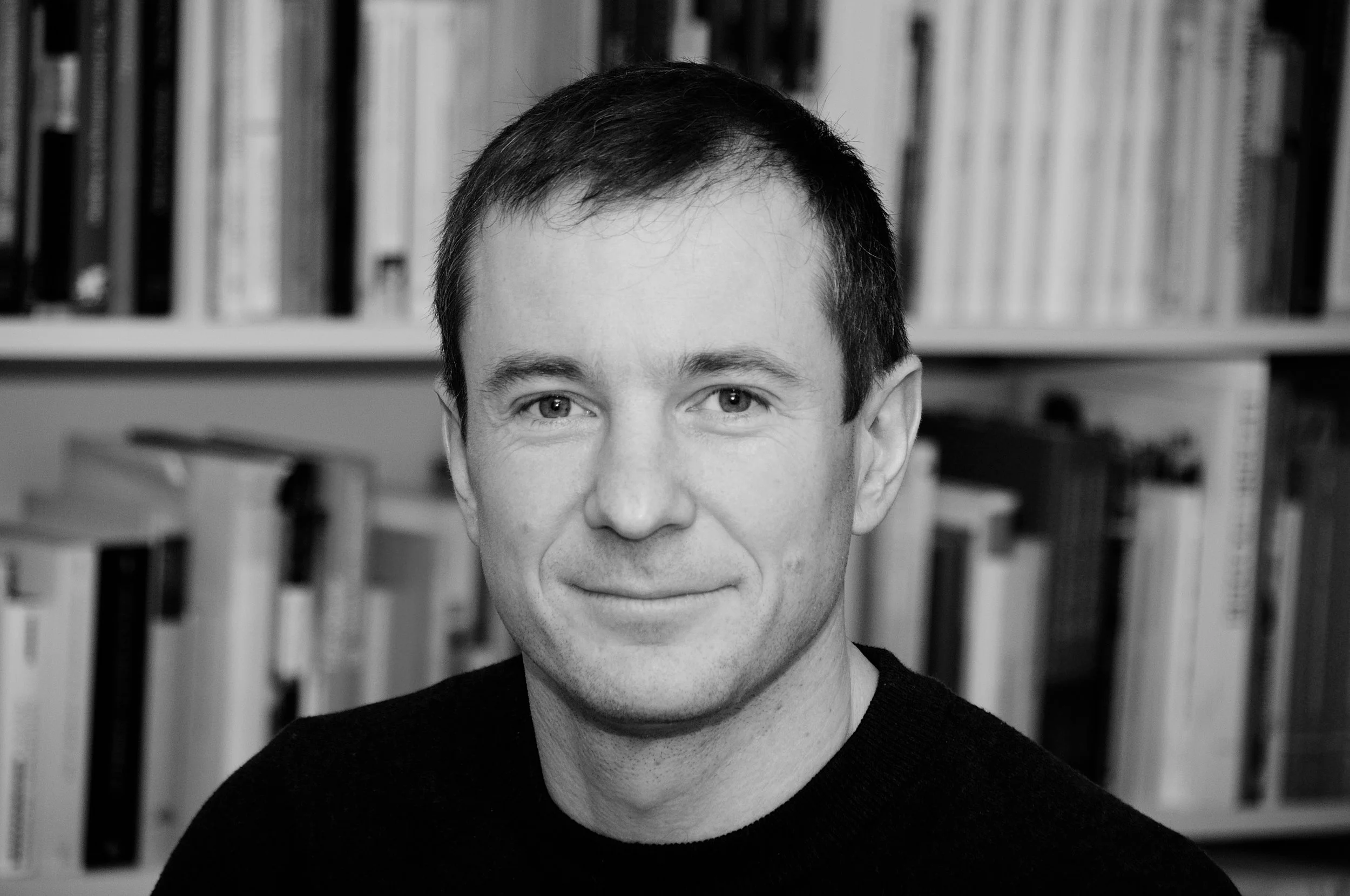 Schwarz-Weiß-Fotografie eines Mannes mit dunklem Haar und freundlichem Ausdruck vor einem Bücherregal.
