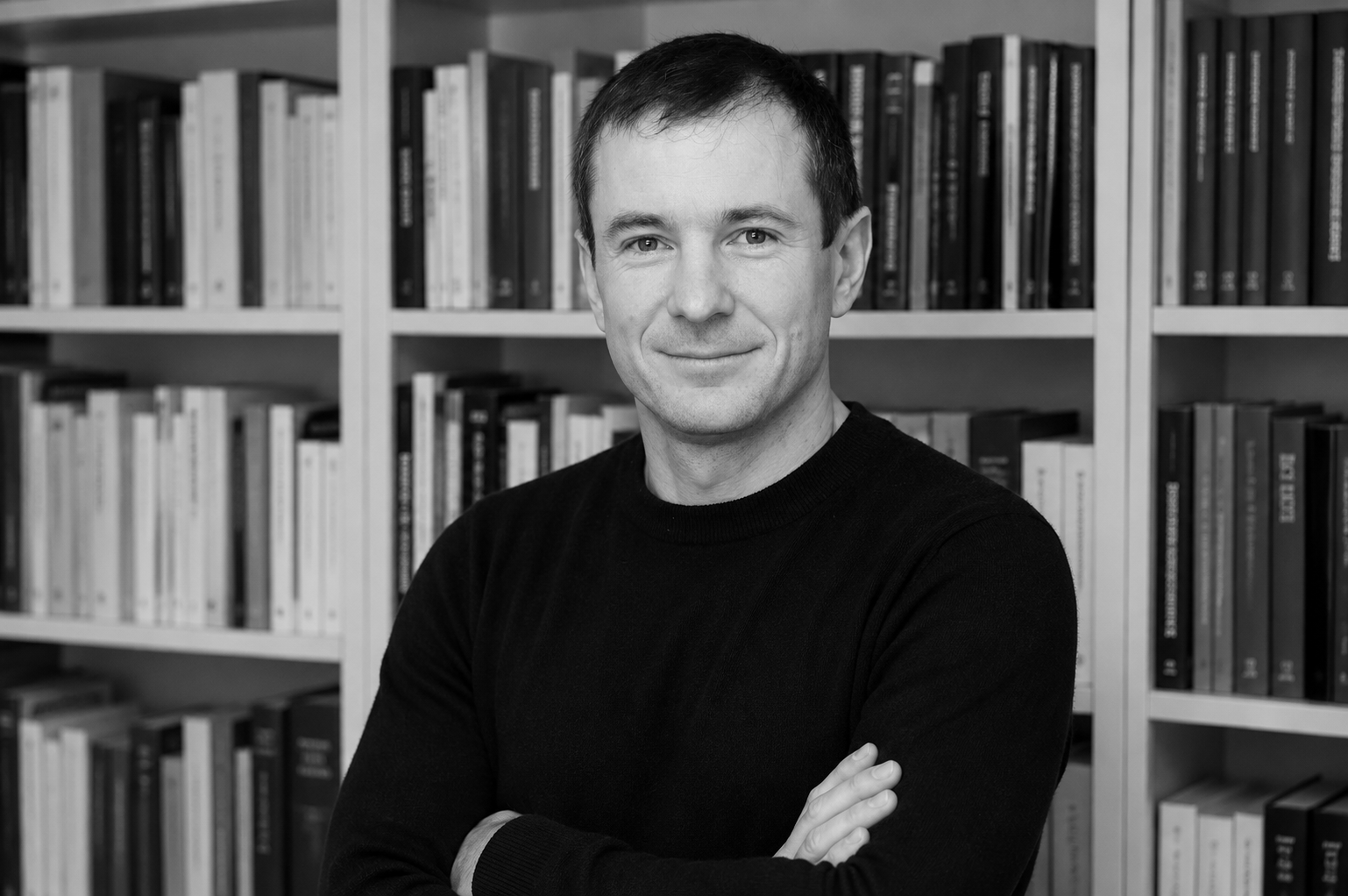 Schwarz-weiß-Fotografie eines Mannes, der vor Büchern steht, mit verschränkten Armen, lächelnd und in einem schwarzen Pullover.