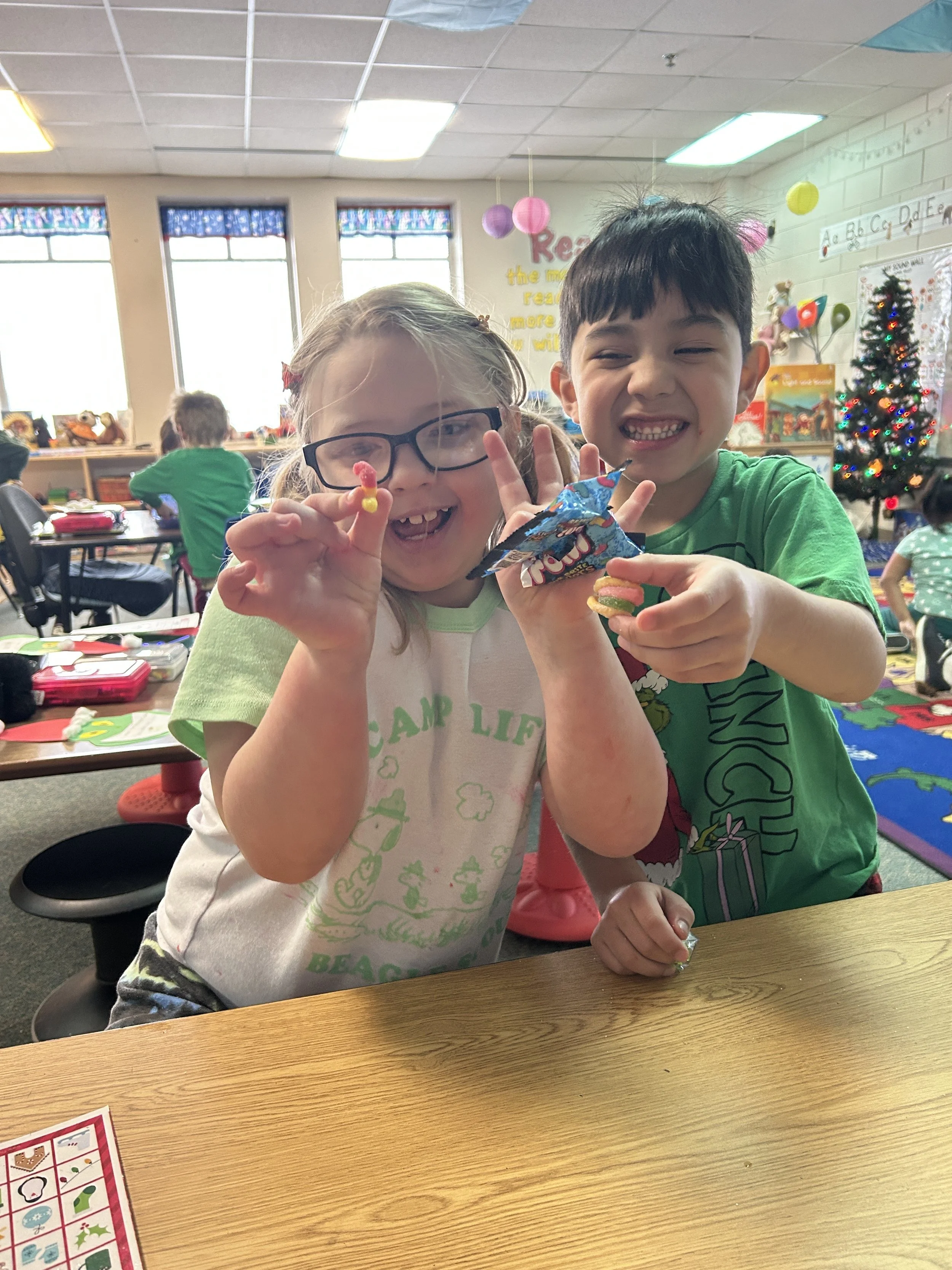 Two children smiling and posing for the camera in a colorful classroom holding candies.