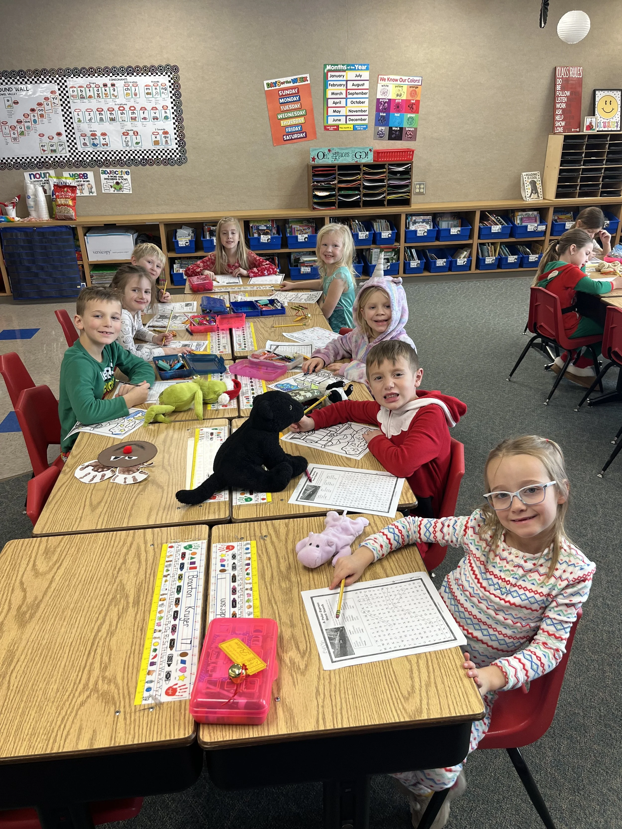 Elementary school classroom with smiling young children sitting at desks engaging in activities with crayons and worksheets. The classroom has decorated bulletin boards, bookshelves, and colorful educational posters on the walls.
