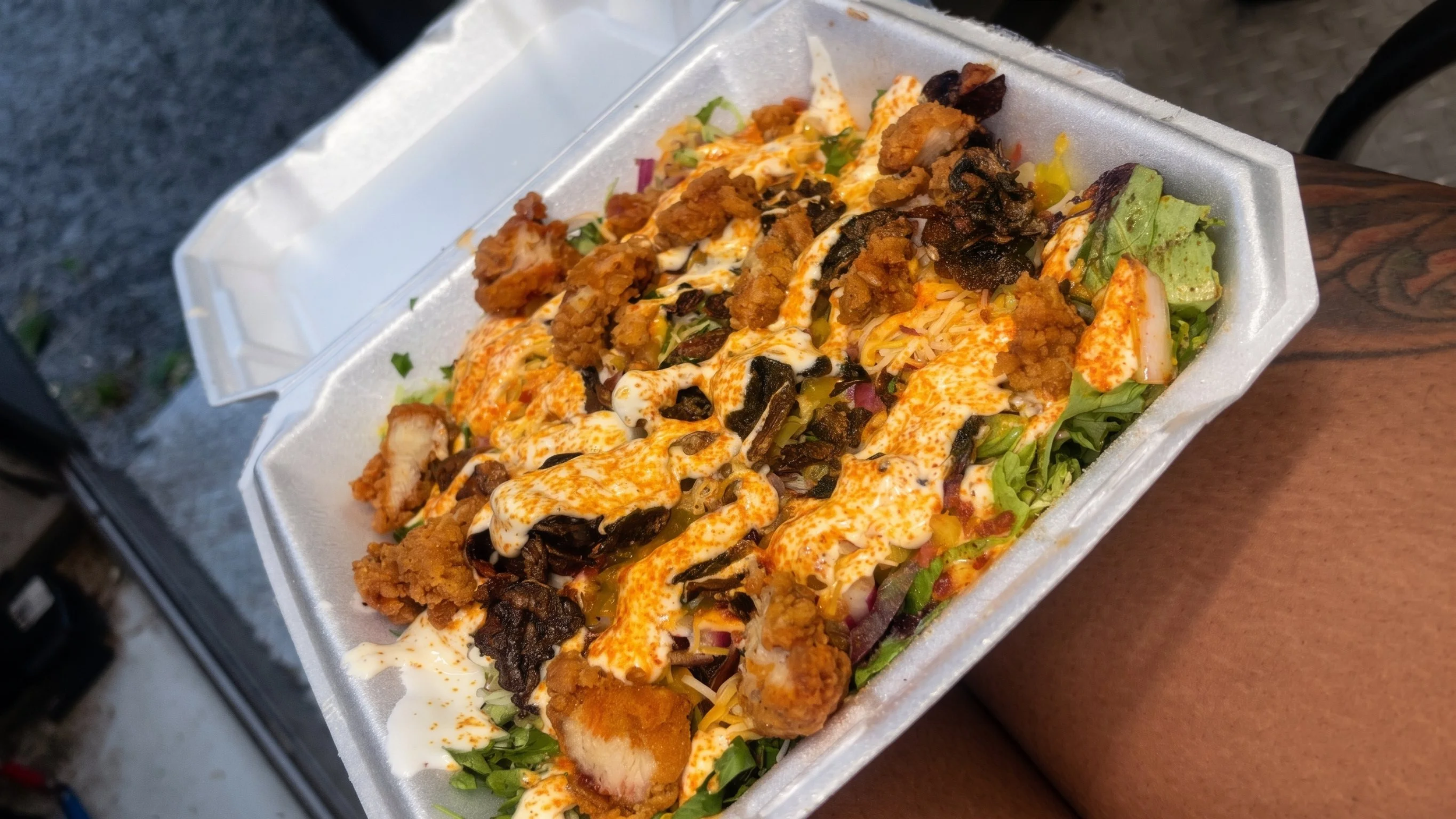 A takeout container filled with a taco salad containing lettuce, fried chicken pieces, shredded cheese, sour cream, melted cheese, diced tomatoes, and black olives.