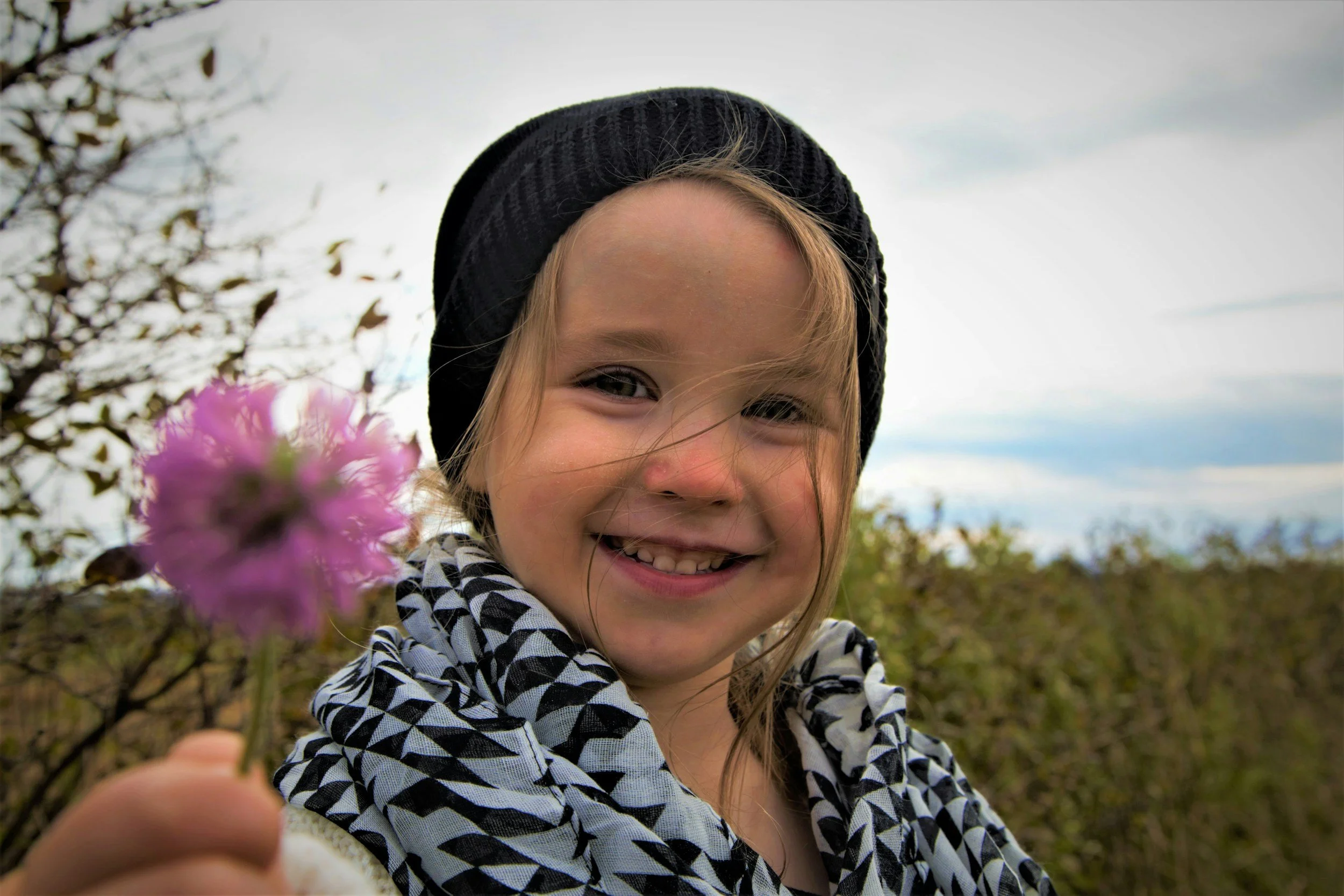 A young girl smiling outdoors, wearing a black knit hat and a black-and-white patterned jacket, holding a pink flower up towards the camera, with a cloudy sky and trees in the background.