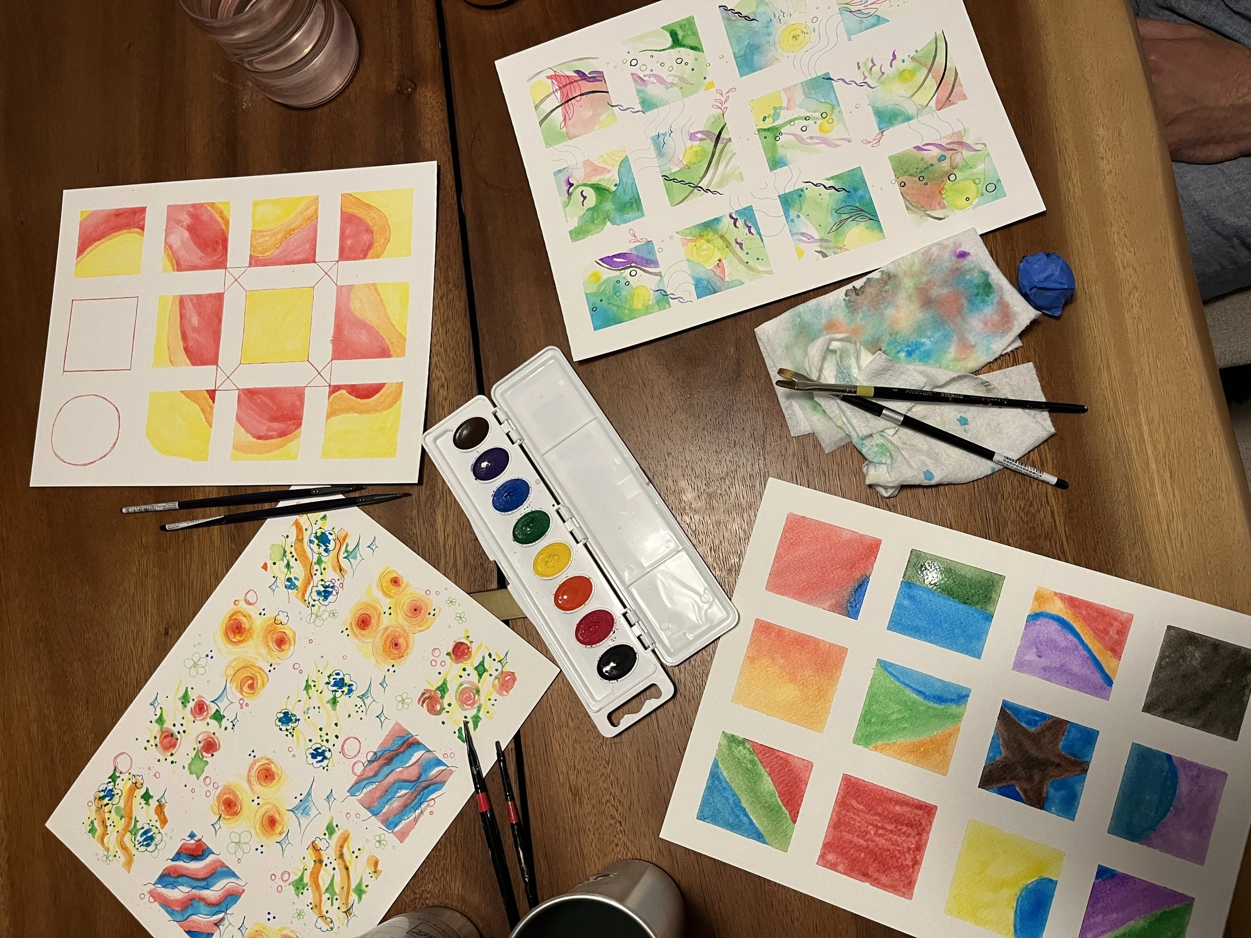 Four watercolor pictures by different artists on a table with watercolors and pens scattered about.