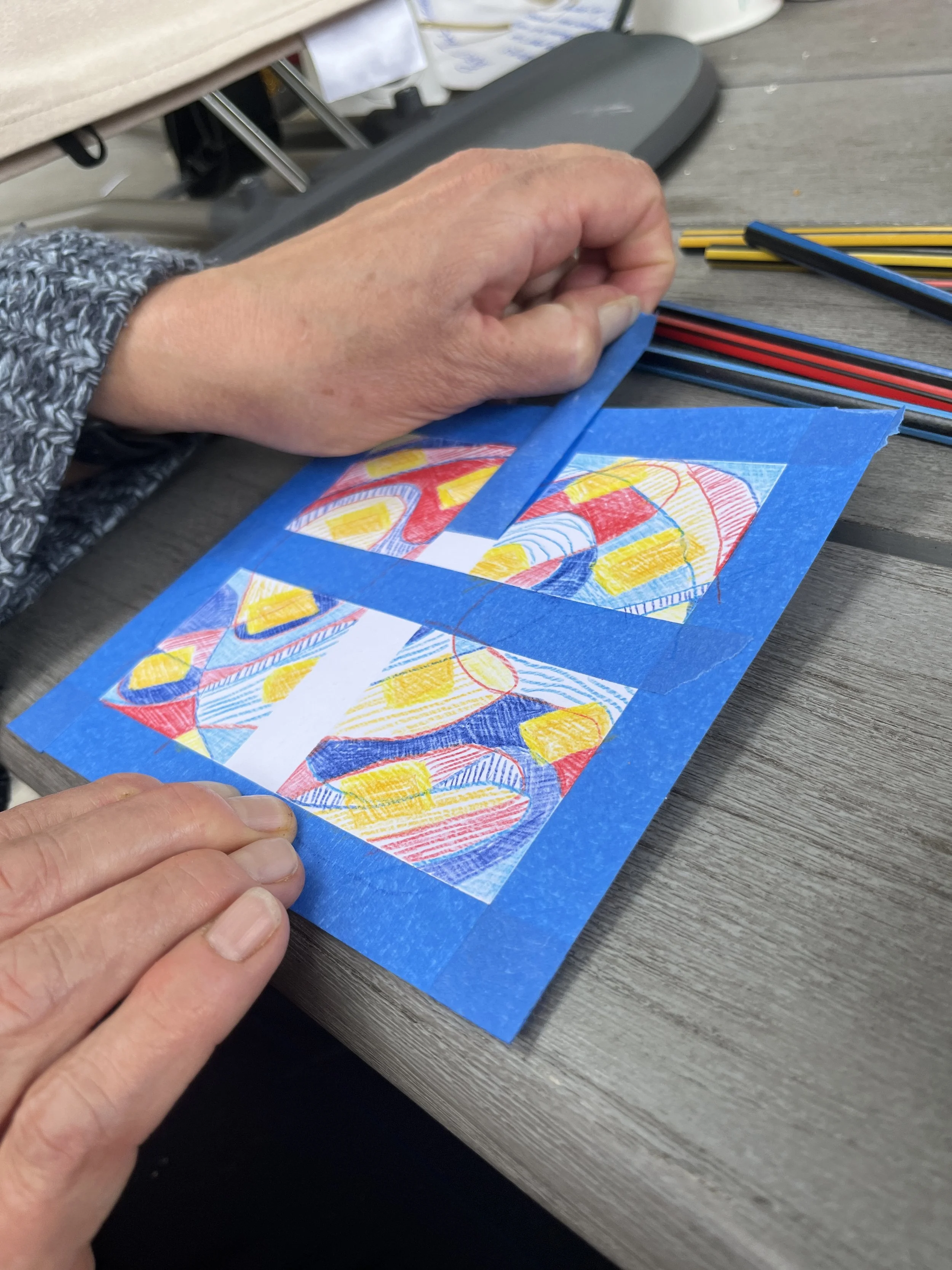square paper with painters tape creating colored in squares. A hand holds the paper while the other hand peels back one piece of tape.