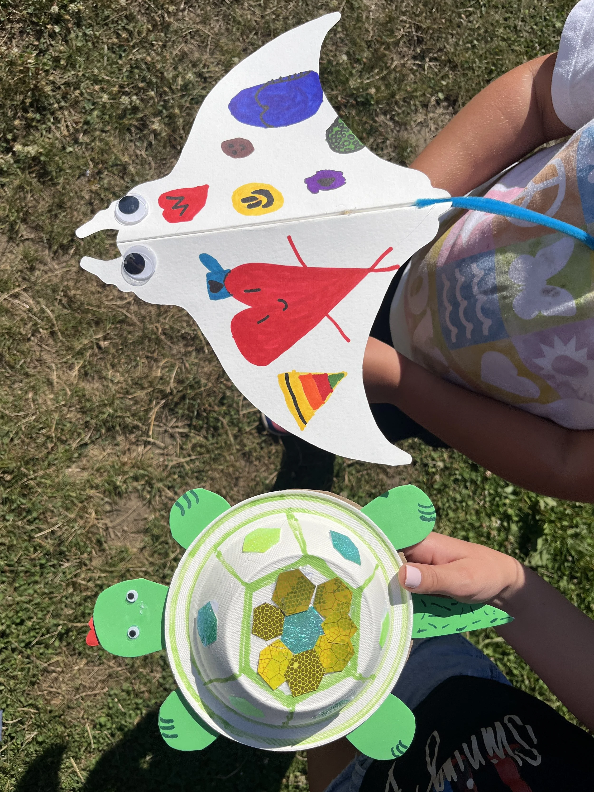 A paper manta ray with drawings and googly eyes and a compostable bowl turtle. Kids showing off their work.