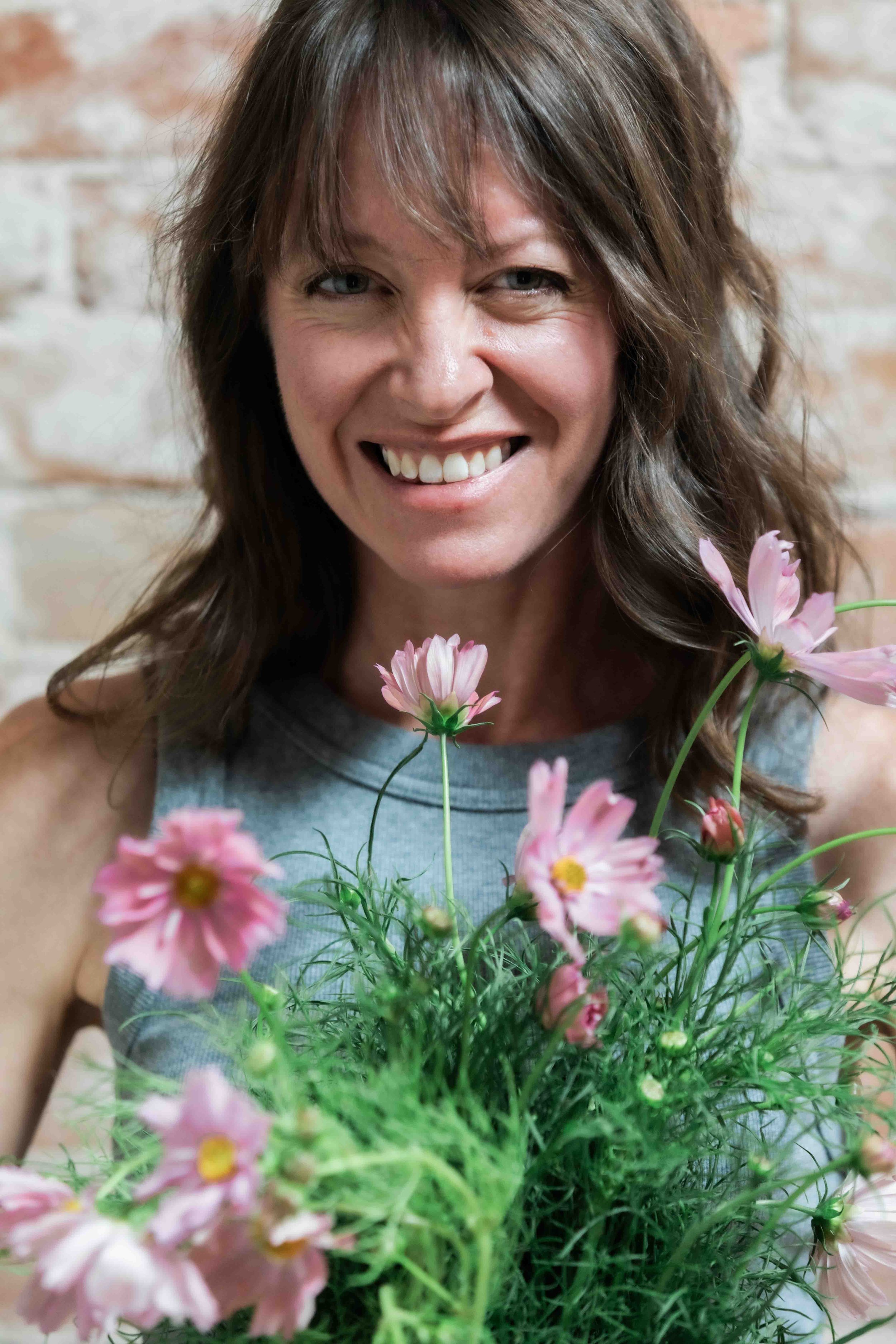 A woman with brown hair holding pink flowers, smiling, in front of a light brick wall.