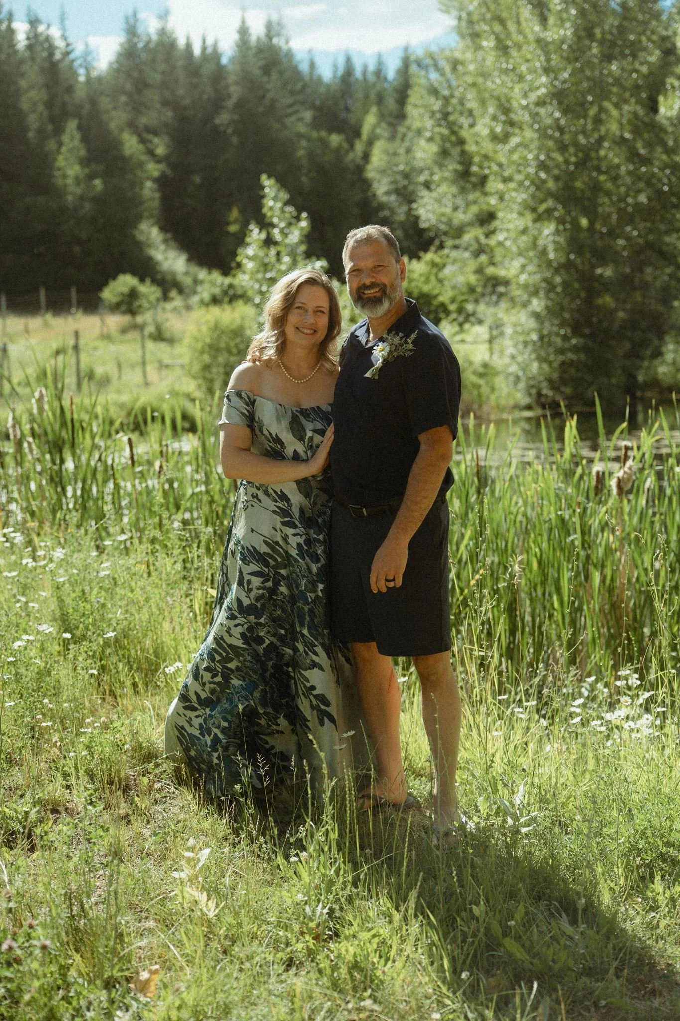 A smiling couple standing in a grassy field near a pond, surrounded by lush green trees on a sunny day.