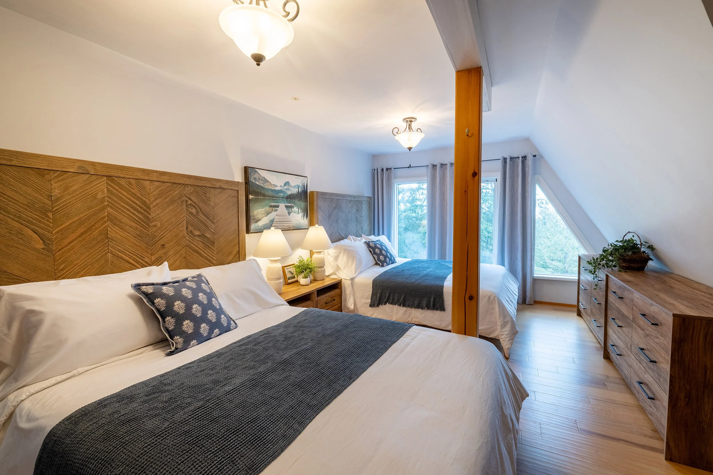 A cozy bedroom with two beds, wooden headboards, and blue and white bedding. There are bedside lamps, a framed landscape painting, and a window with curtains revealing trees outside. A wooden dresser is on the right, and a wooden support beam runs through the room.