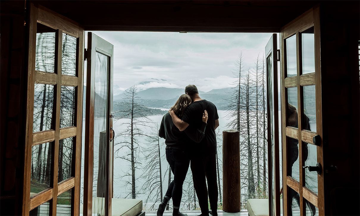 A couple embraces on a balcony with a view of a misty lake, mountains, and trees in the background.