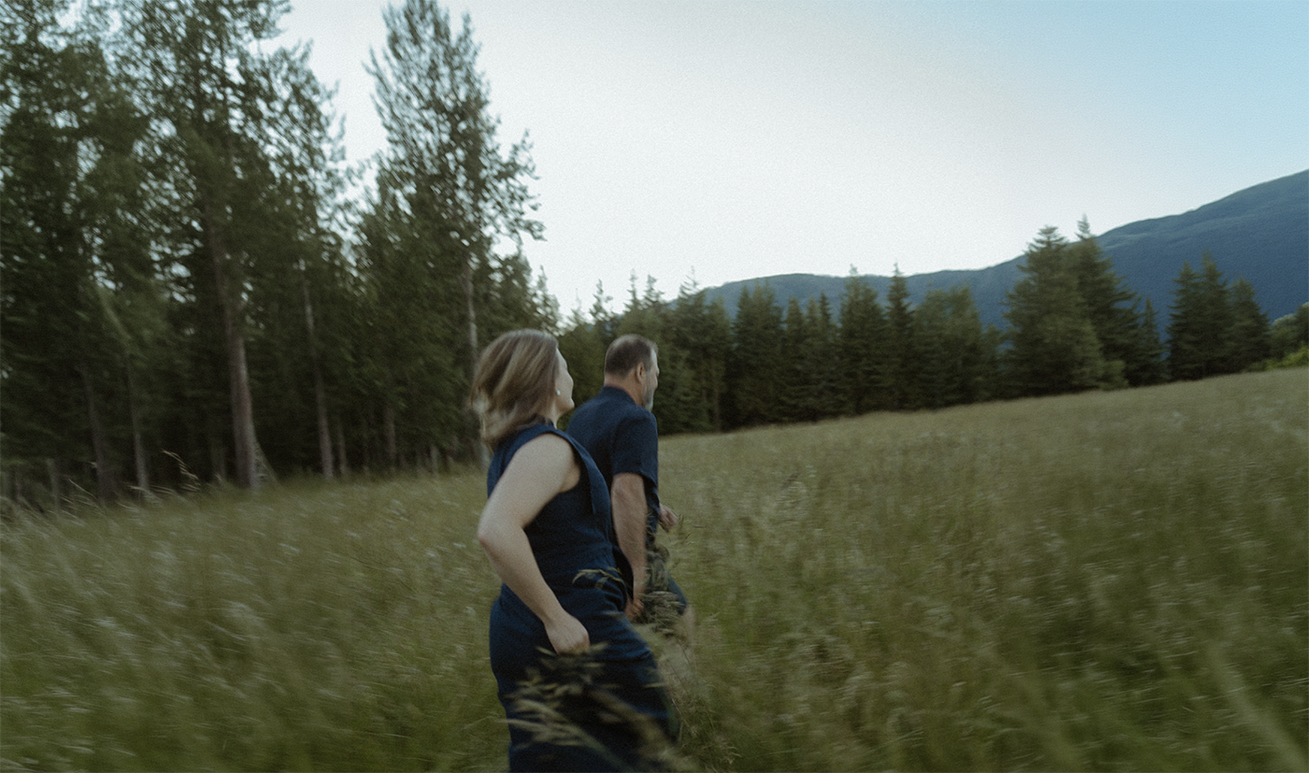 A man and a woman hiking through a grassy field with trees and mountains in the background.