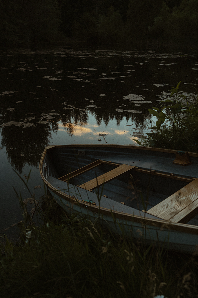 A small boat docked at the edge of a calm, dark lake surrounded by trees during twilight.
