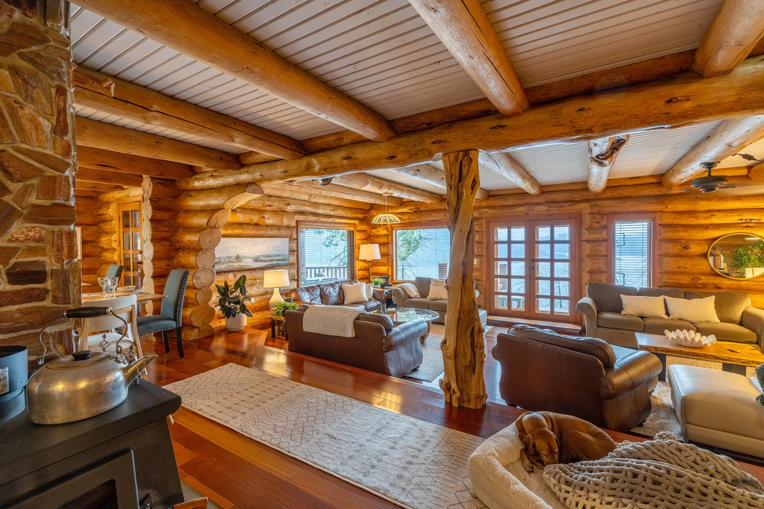 Interior of a rustic log cabin living room with wooden beams, log walls, large windows, cozy seating, and warm lighting.