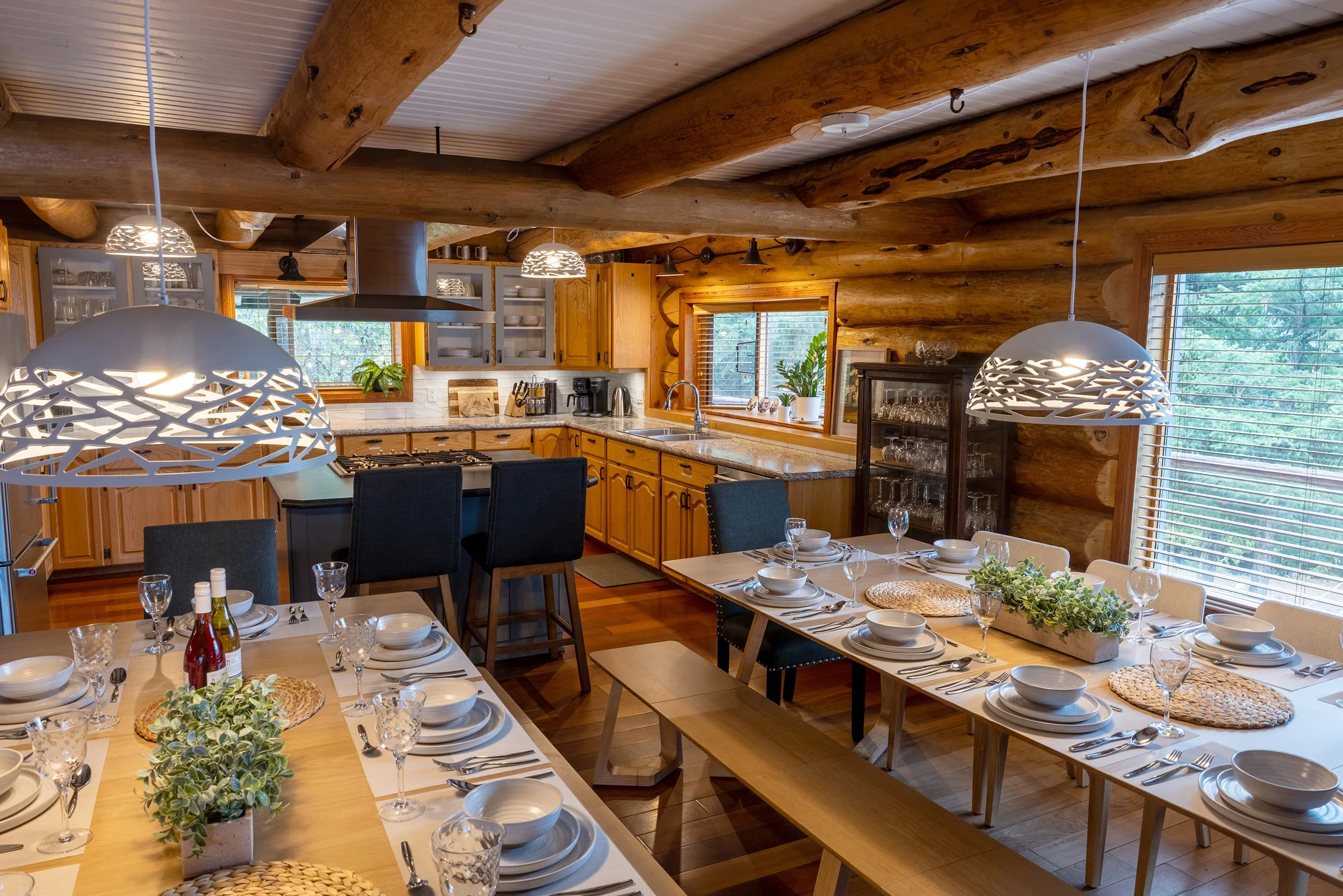 Interior of a rustic kitchen and dining area with wooden beams, cabinets, and large windows. The table is set with white dishes, glasses, and a decorative centerpiece, indicating preparation for a meal or gathering.