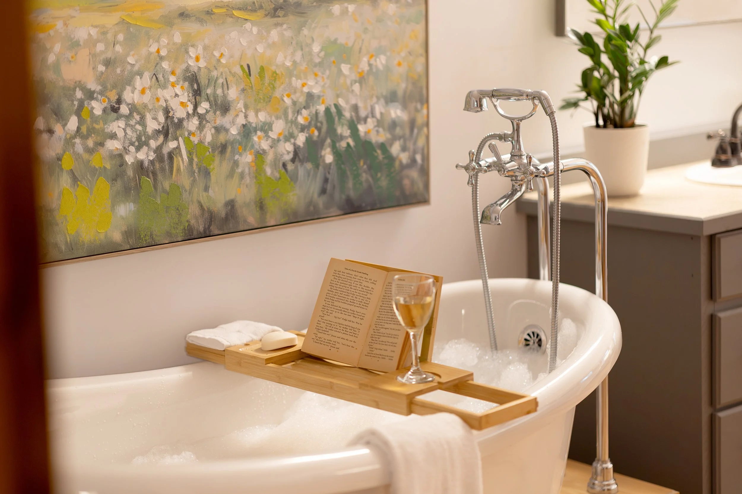 A cozy bathtub scene with a bubble-filled bathtub. A wooden tray holds an open book, a glass of wine, soap, and towels. A chrome faucet and a potted plant are visible in the background, with a colorful abstract painting on the wall.