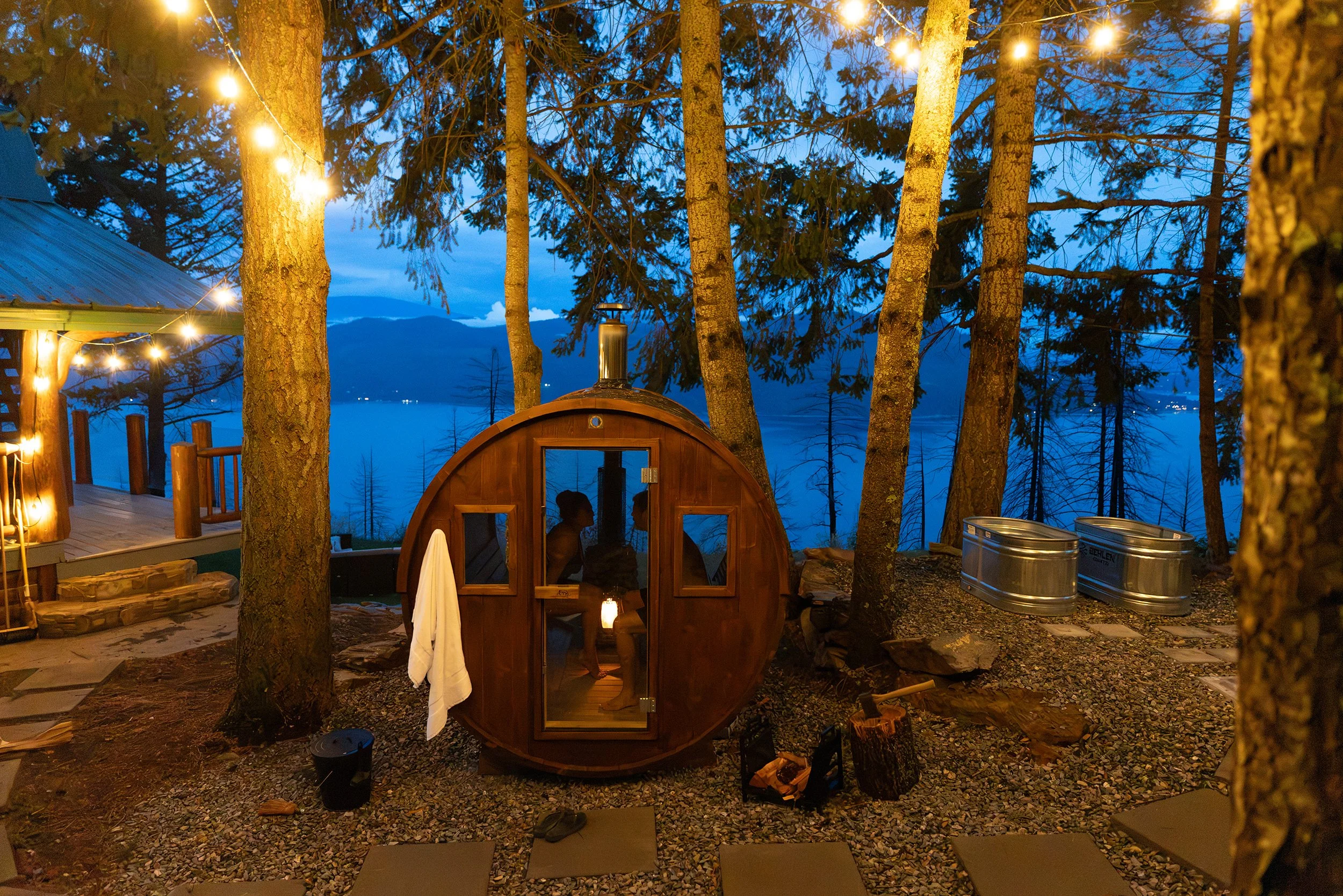 An outdoor scene at twilight with string lights hanging among tall trees overlooking a lake or river. There is a wooden sauna or small building with a chimney, and people are visible through its windows. The area is decorated with patio furniture and