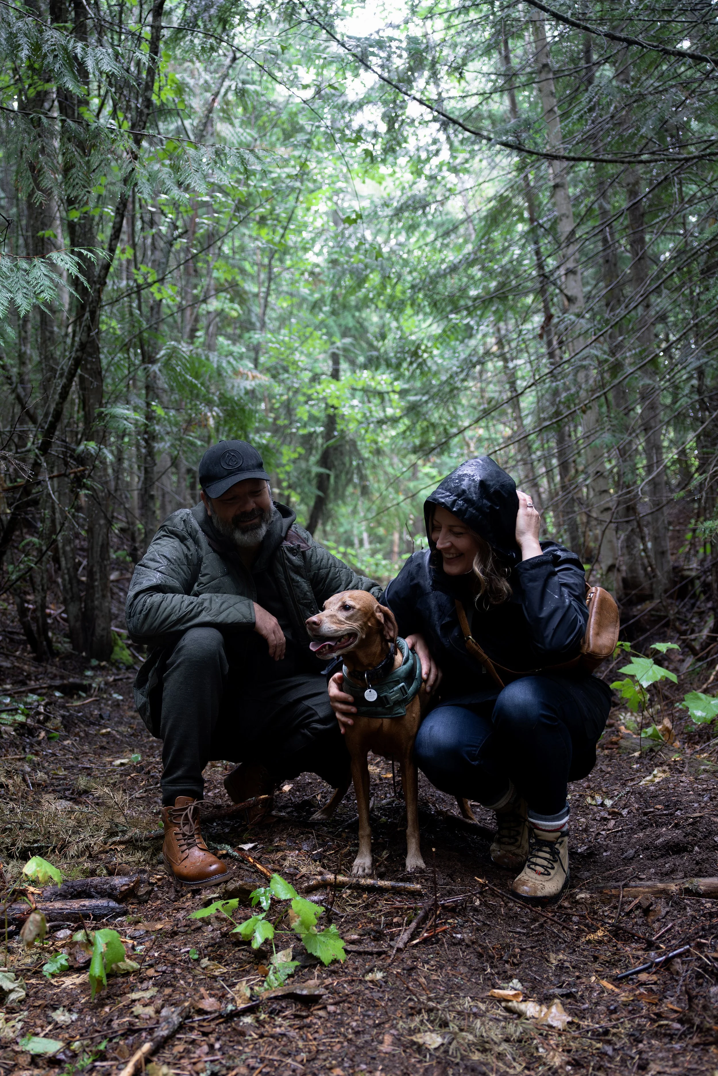 Two people, one man and one woman, are in a forest with their dog. They are squatting on the ground, smiling and petting the dog. The man has a beard and is wearing a cap and outdoor jacket. The woman has a hooded jacket and is touching her hair. The